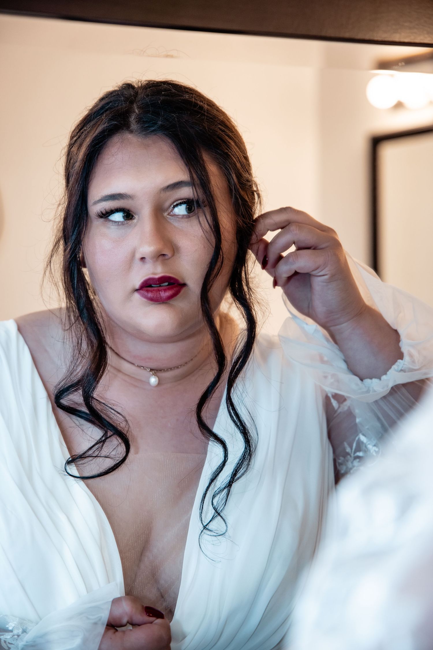 Bride with dark hair and red lips adjusting her hair while getting ready in white bridal robe.