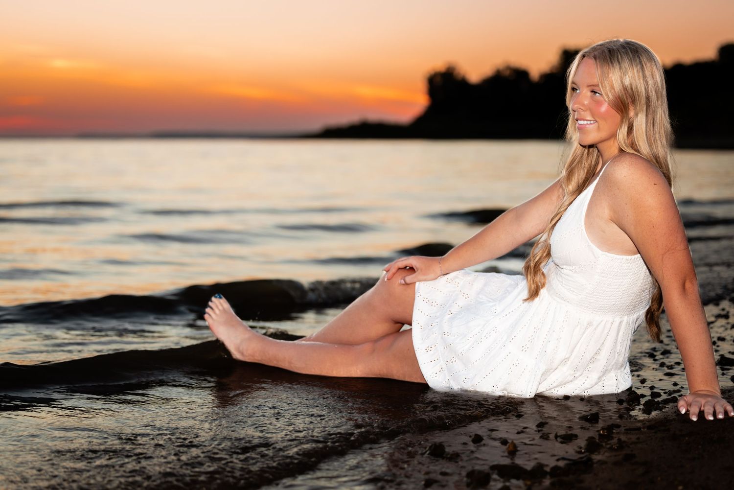 Blonde woman in white dress sitting on rocky beach at sunset with golden sky reflecting on calm water.