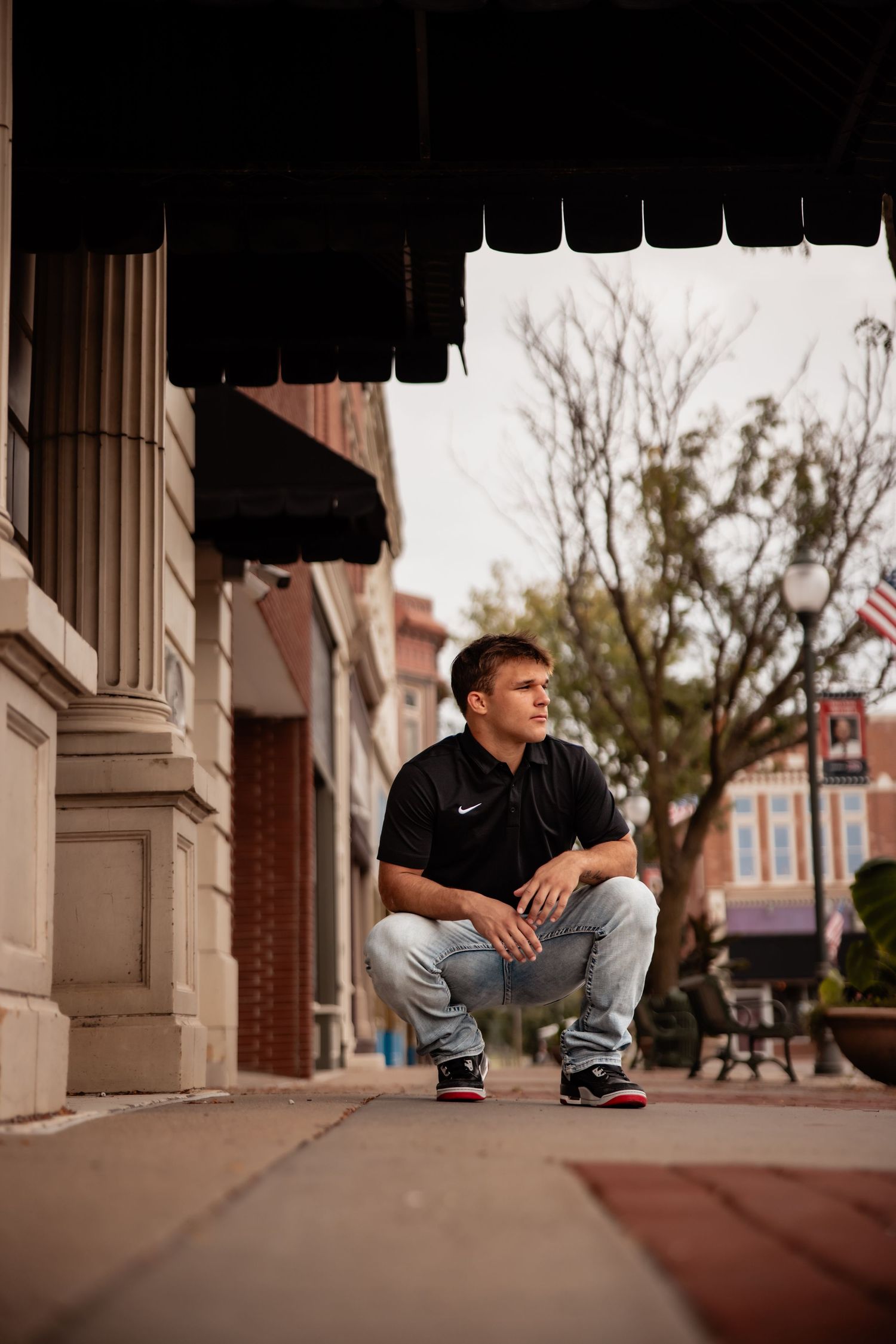 Young man in black Nike polo and jeans crouching on urban sidewalk under awning, gazing thoughtfully into distance.