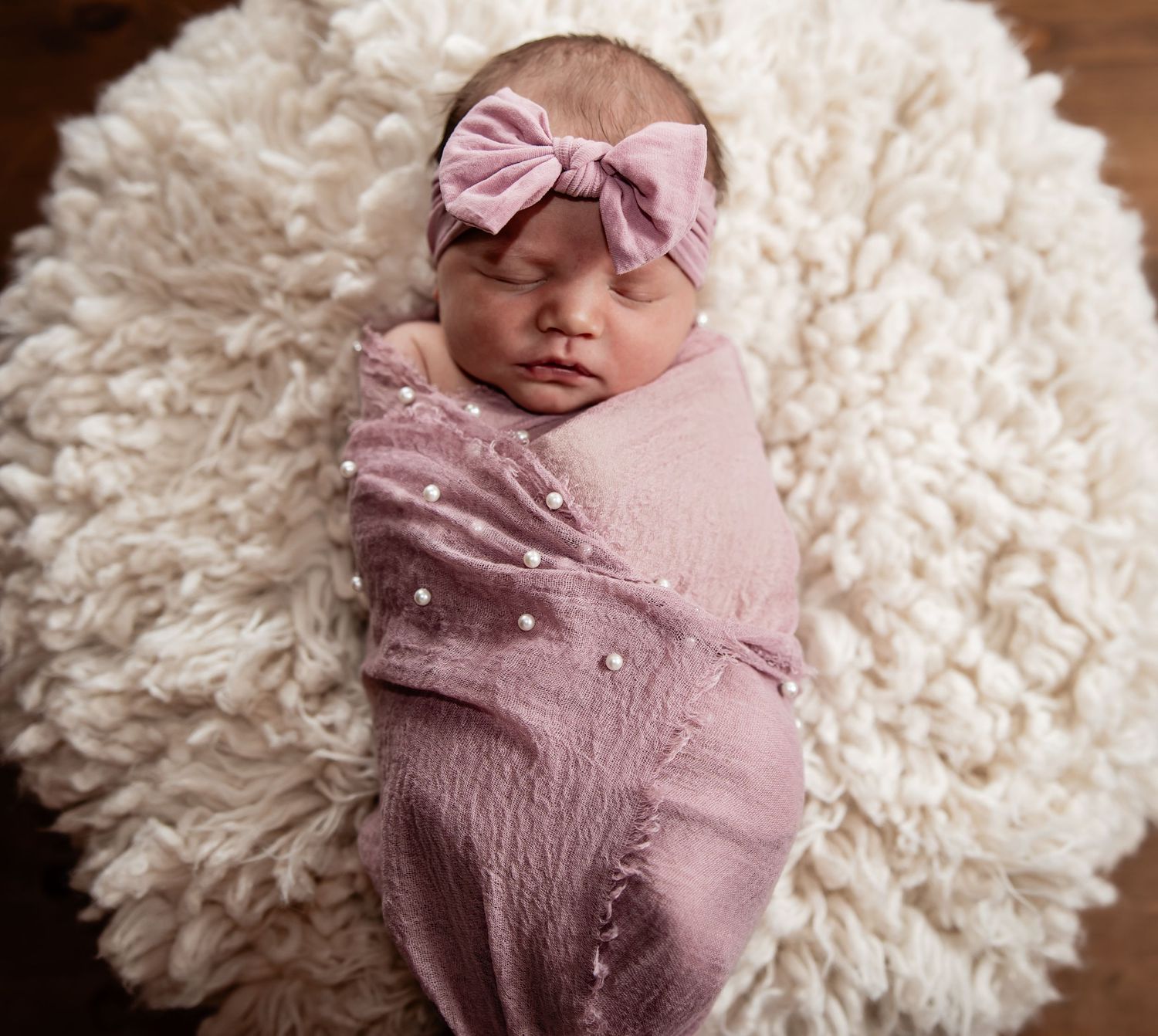 Newborn baby girl swaddled in pink wrap with pearl accents, wearing a pink bow headband, resting on fluffy white backdrop.