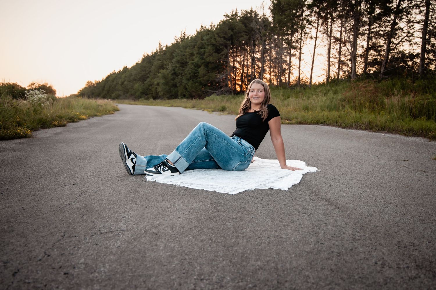 Young woman in jeans and black top sitting on a white blanket on a road at sunset with trees in background.
