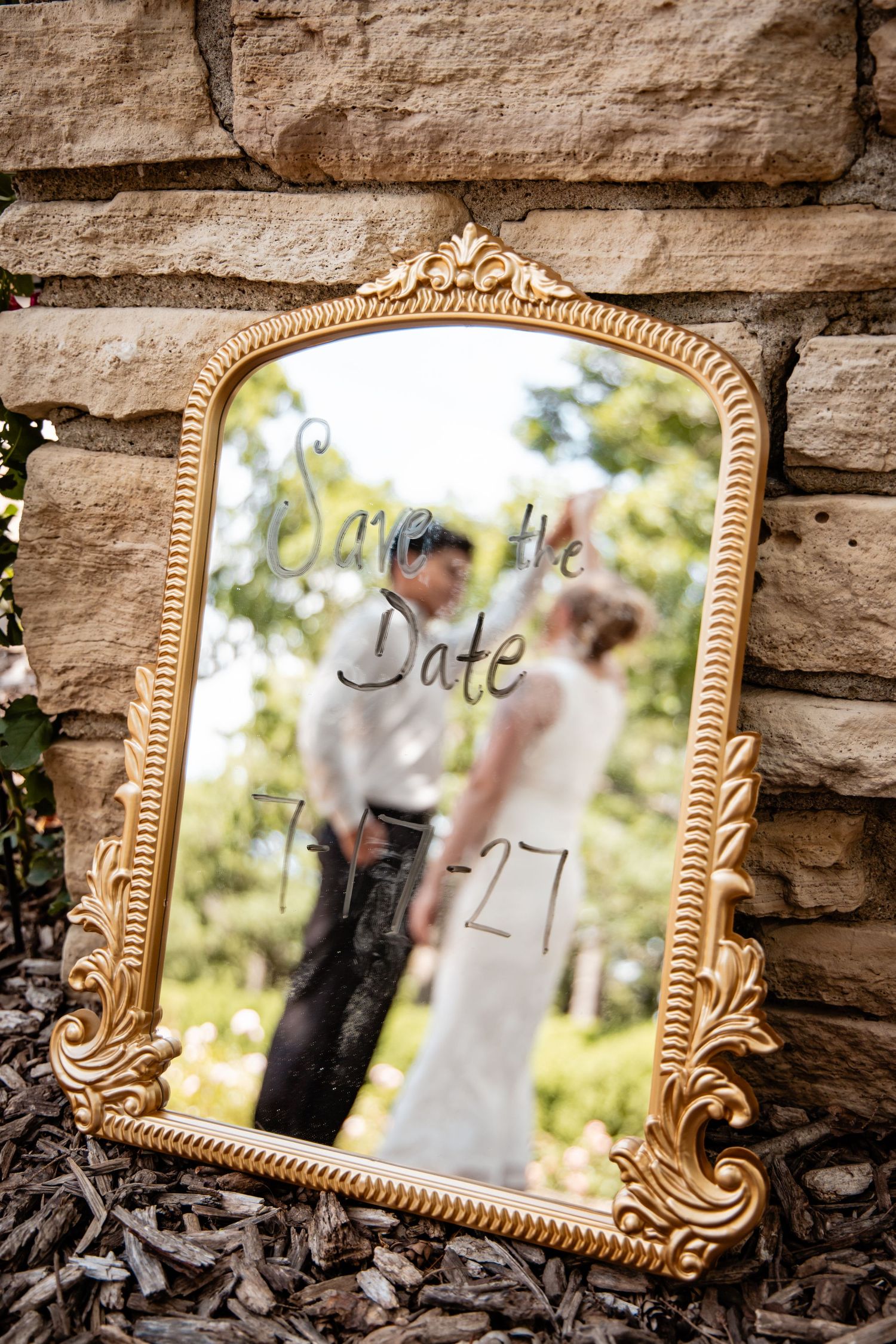 Gold ornate mirror with Save the Date 7-27 written on it, reflecting a bride and groom outdoors.