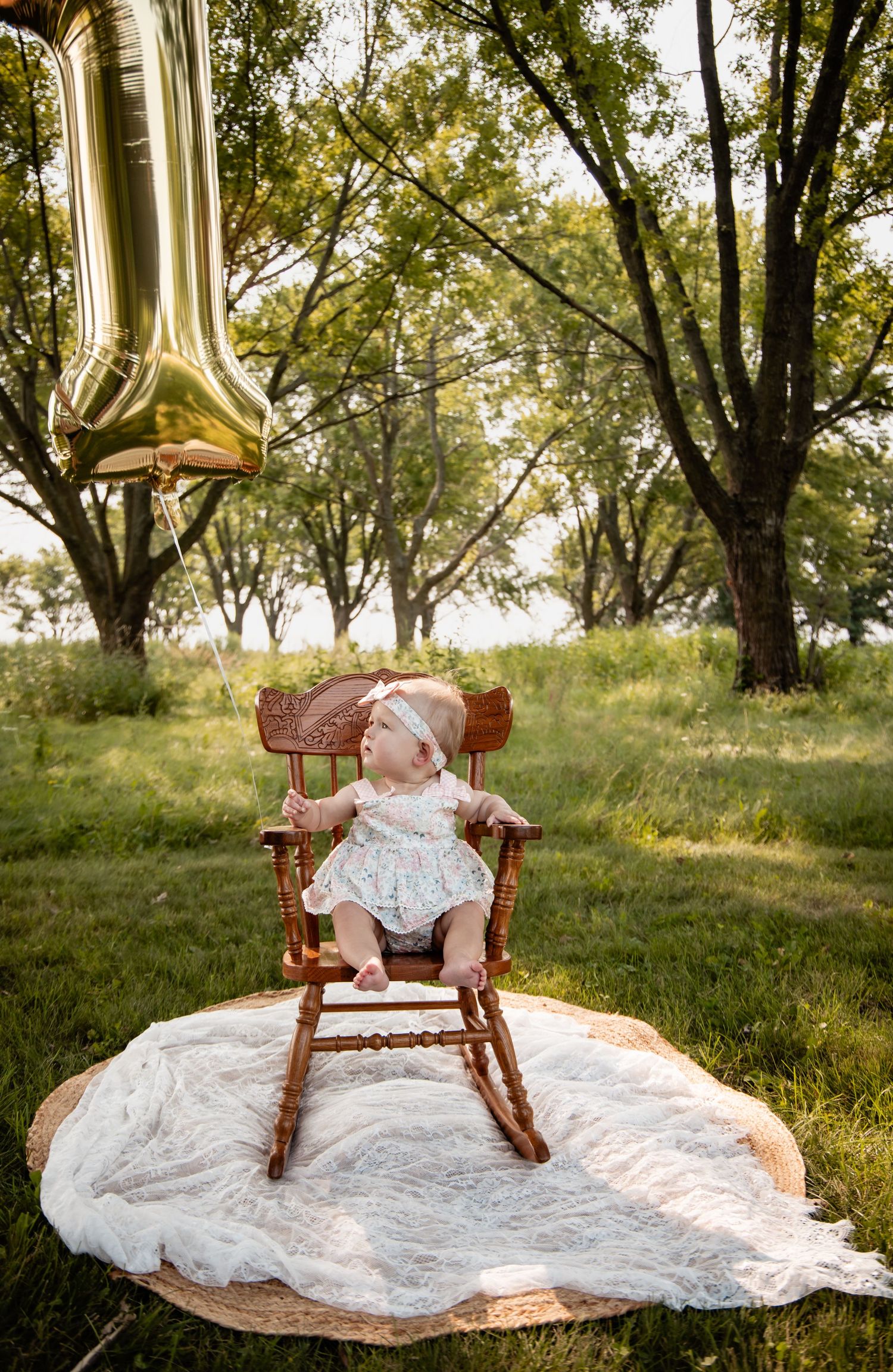 Baby girl in floral romper sits in wooden rocking chair outdoors with gold number 1 balloon for first birthday.