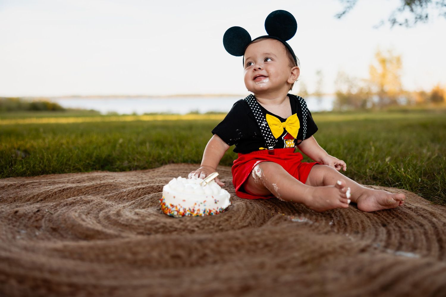 Baby in Mickey Mouse costume sits on sand near a smash cake outdoors during golden hour.