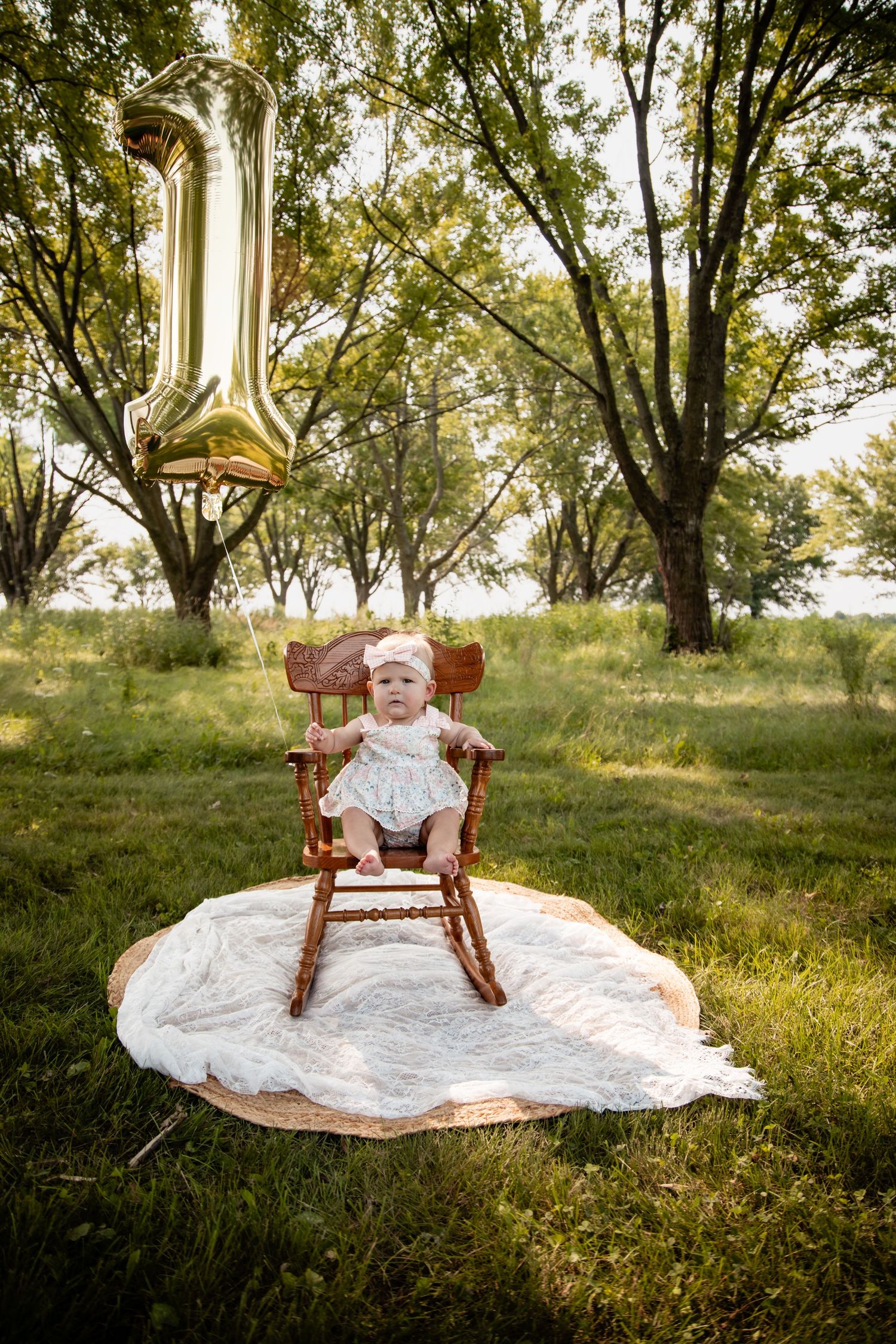 Baby girl in white dress sits in wooden rocking chair outdoors with gold number 1 balloon for first birthday celebration.