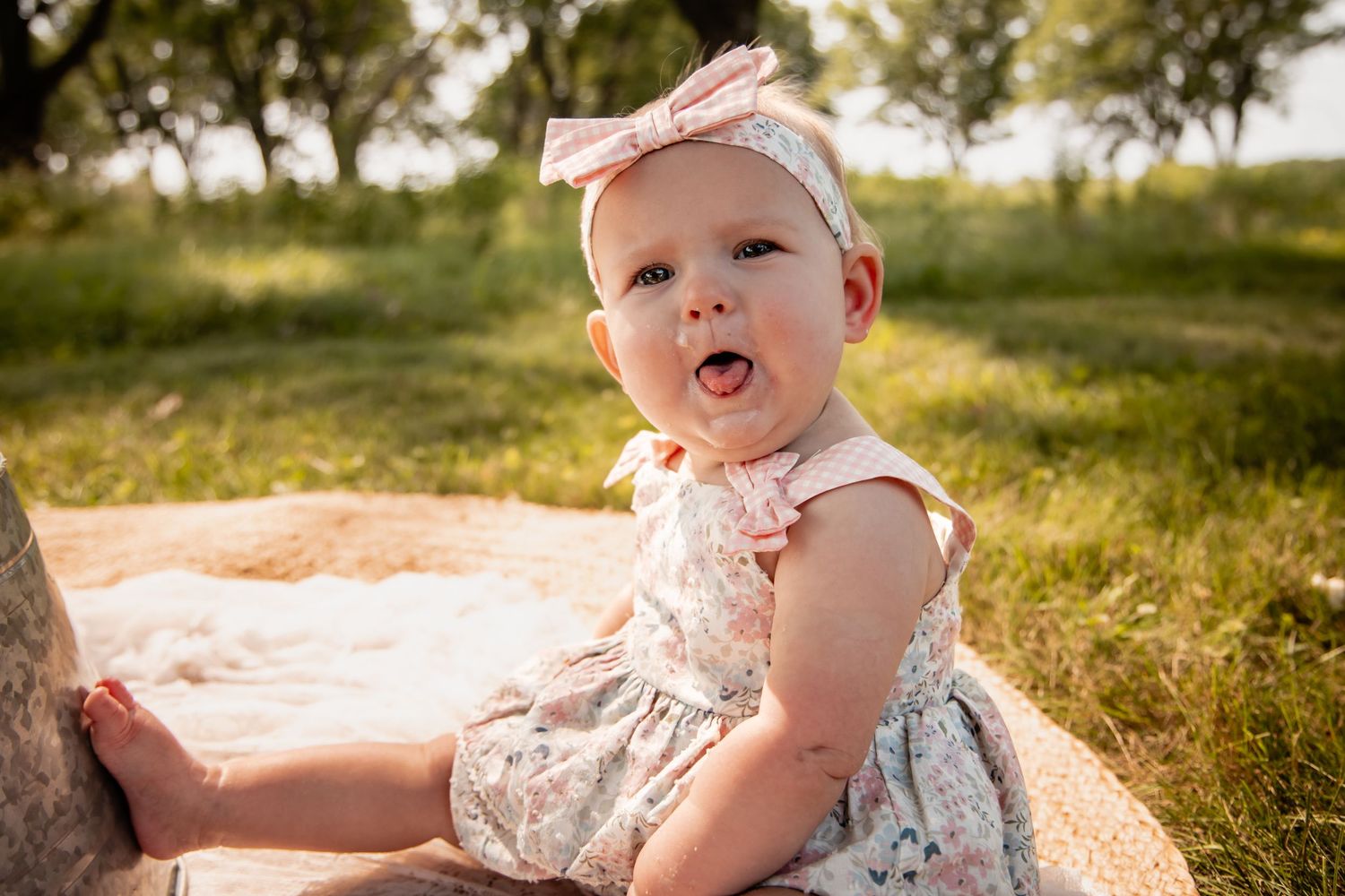 Adorable baby girl in floral romper and pink bow headband sitting outdoors on blanket in sunny green field.