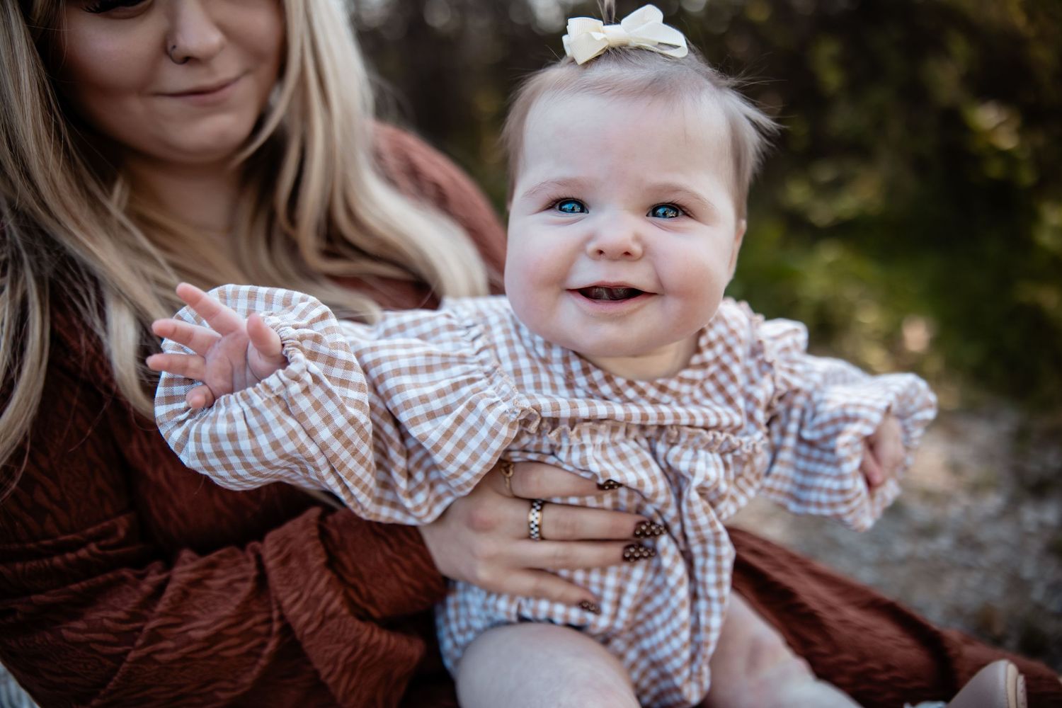 A happy baby girl in a plaid dress with a white bow smiles while being held by her mother outdoors.