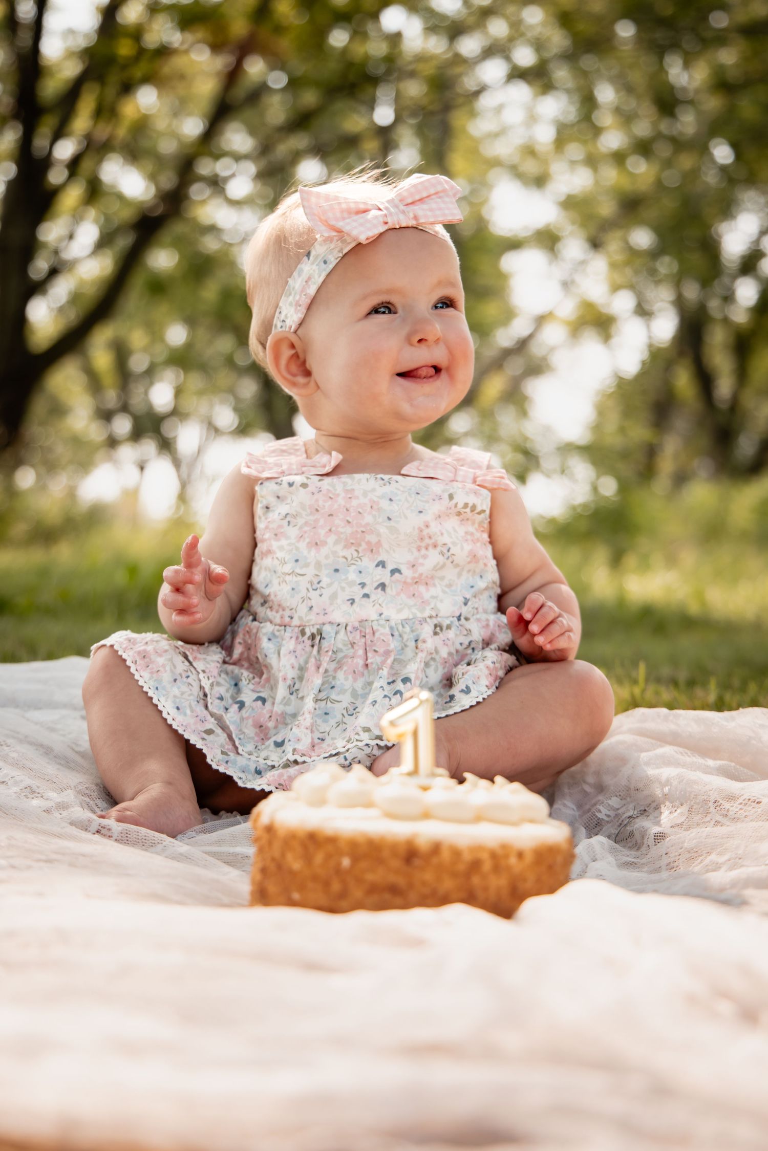 Baby girl in floral romper celebrates first birthday with cake outdoors on blanket in sunny park setting.