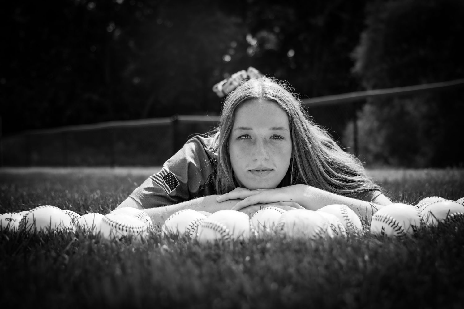 Black and white photo of a girl lying on grass resting her chin on softballs outdoors.