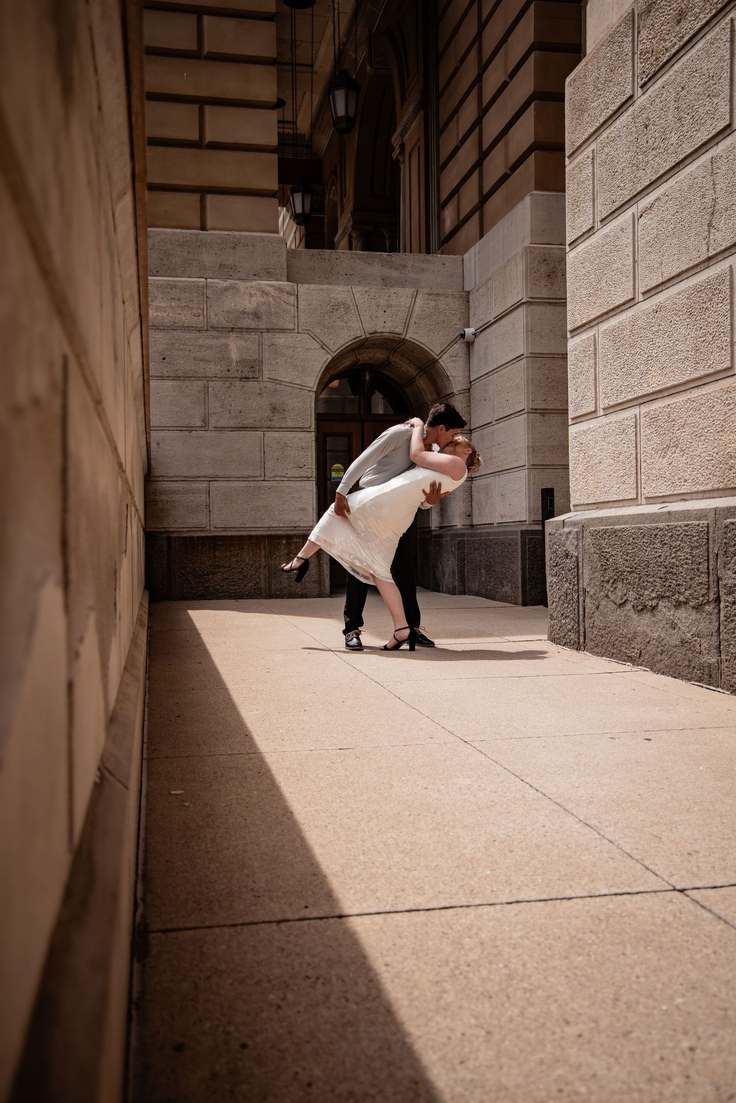 Couple sharing a romantic dip kiss in a sunlit stone building corridor, bride in white dress, groom in gray suit.