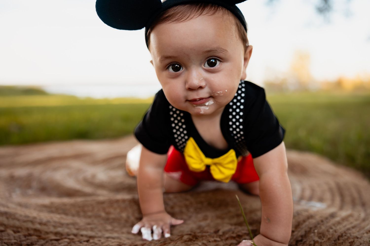 Adorable baby in Mickey Mouse costume crawling on ground outdoors during golden hour photo shoot.