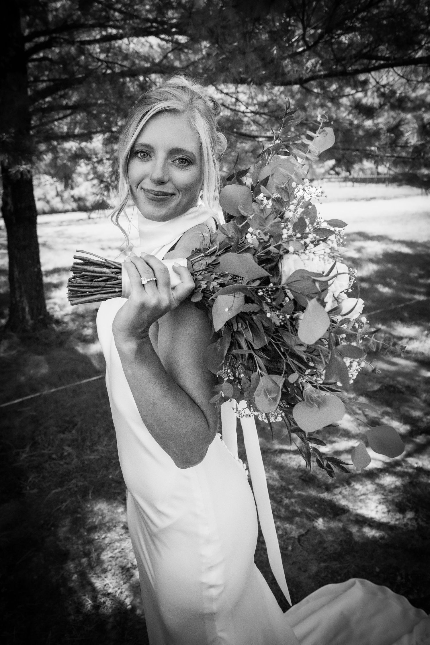 Black and white photo of a smiling bride in a backless gown holding a lush eucalyptus bouquet outdoors.