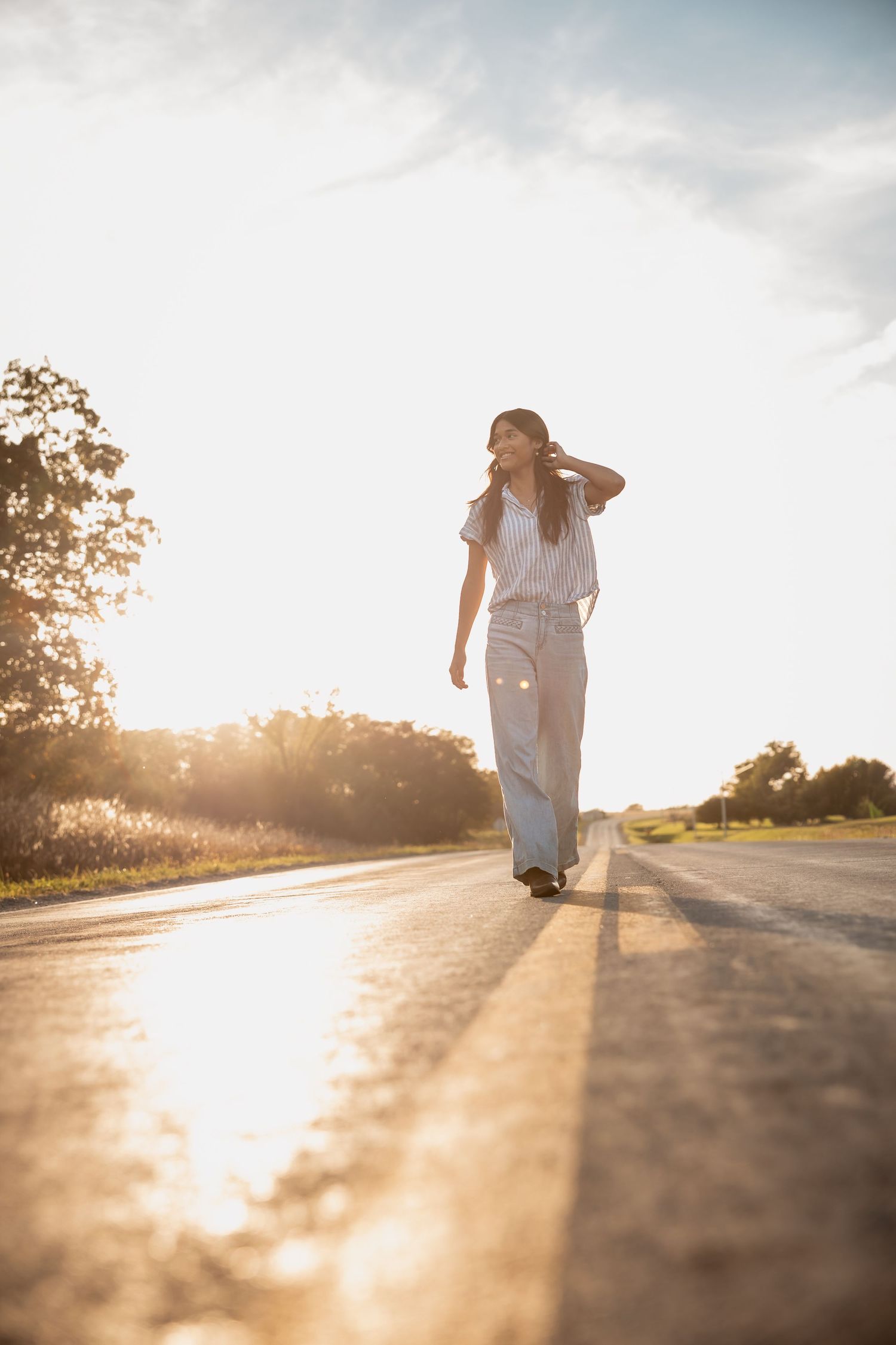 Woman in white top and wide-leg pants walking on a sunlit road during golden hour.