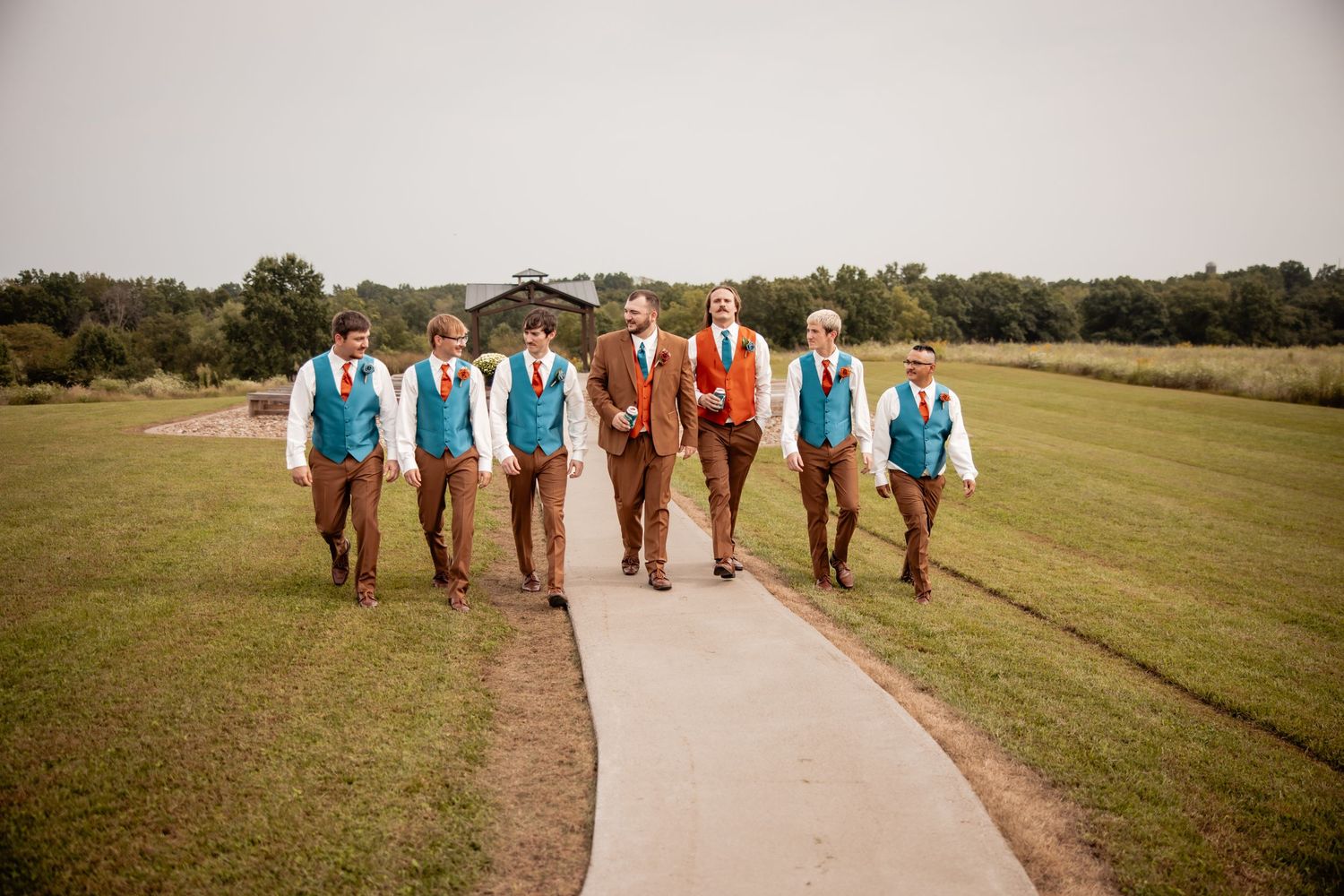 Groom in rust suit walks with groomsmen in teal vests and brown pants on a path through green fields.