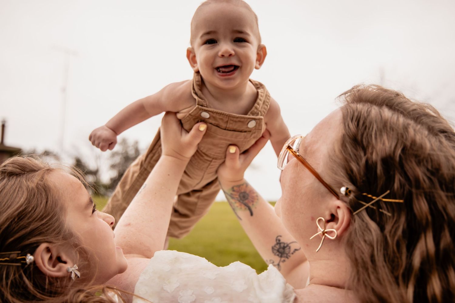 Two women joyfully lift a laughing baby in brown overalls outdoors, sharing a heartwarming family moment.
