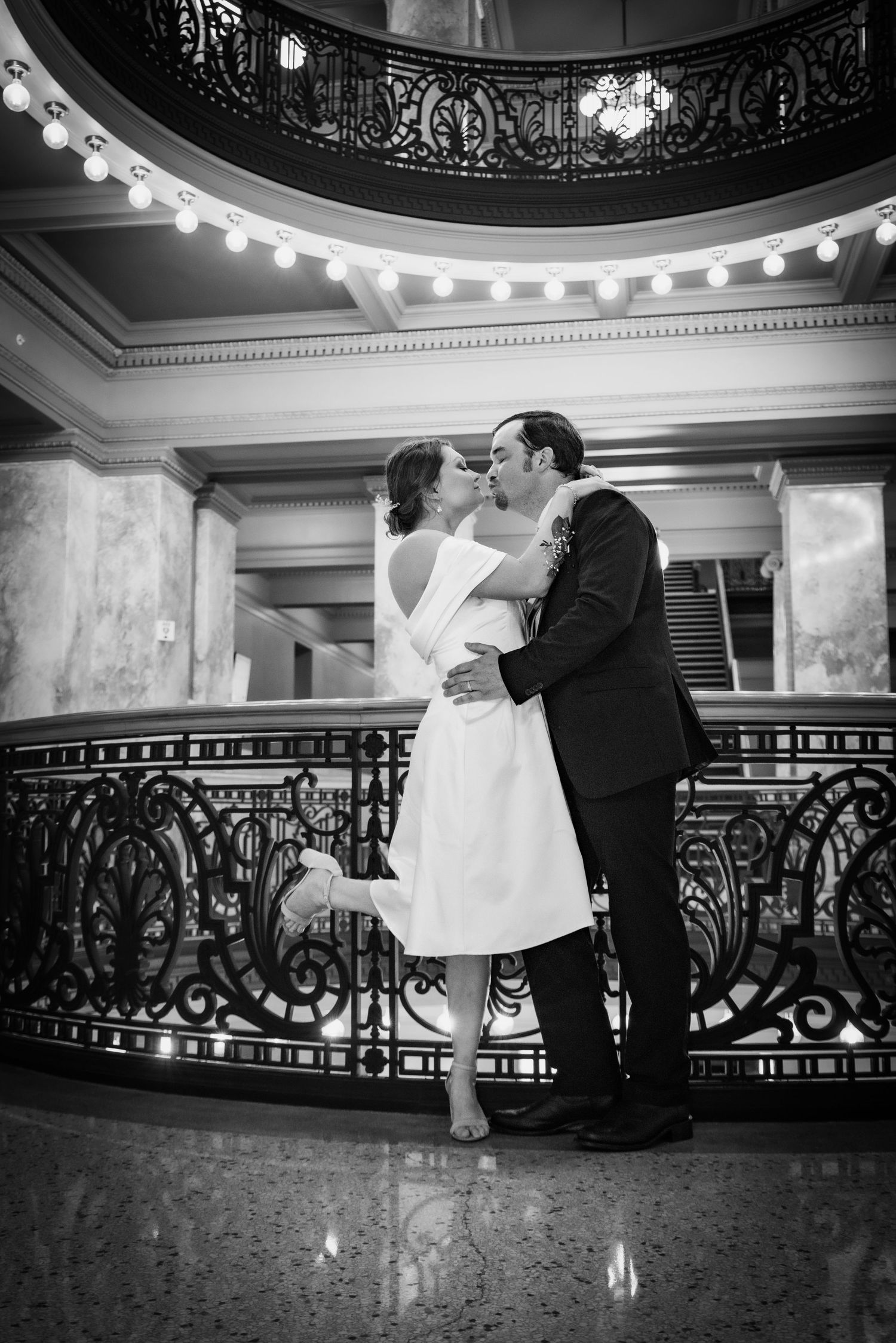 Black and white photo of a couple sharing a romantic kiss in an elegant ballroom with ornate iron railings.