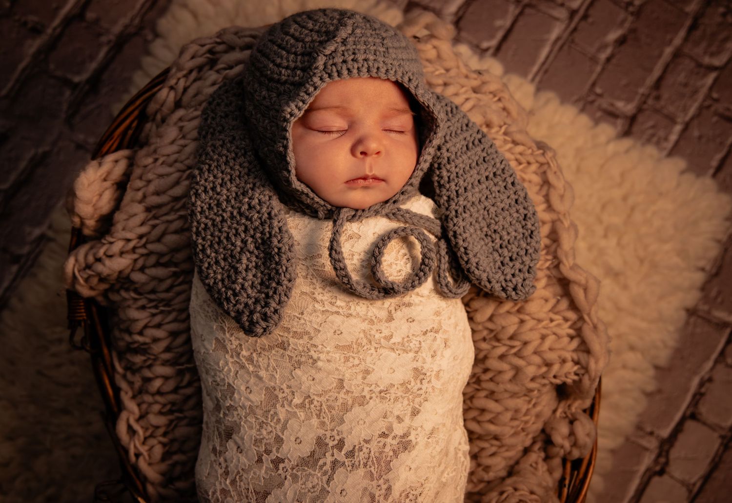 Sleeping newborn baby wrapped in gray crochet knit swaddle with bunny ear hat, nestled in a wicker basket.