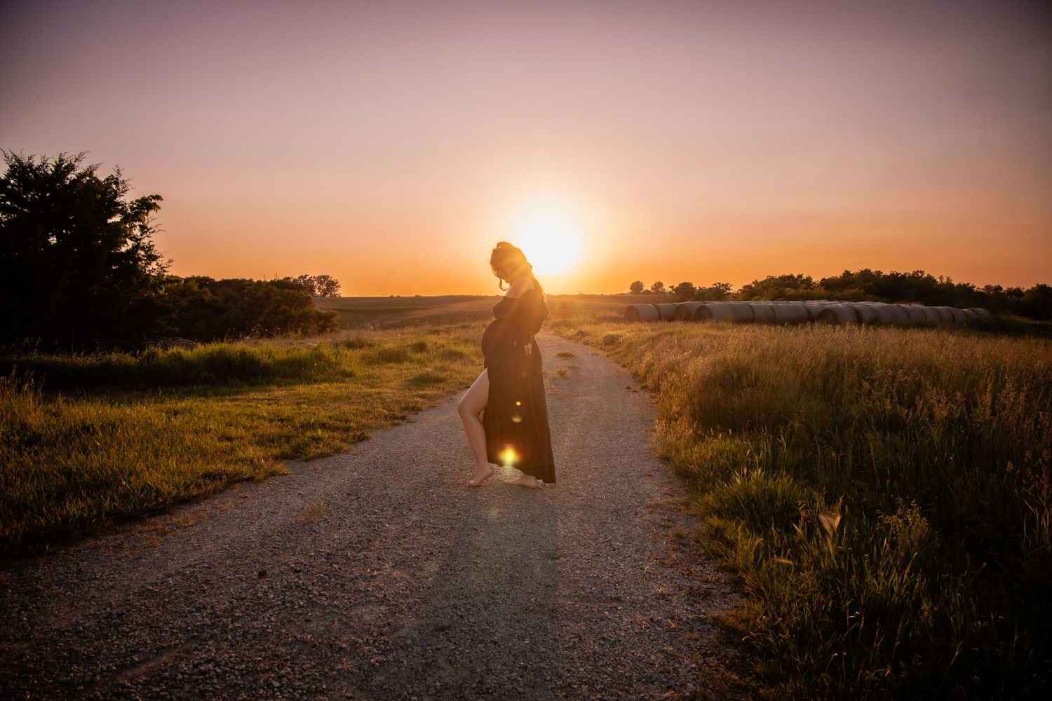 A woman in a dark dress stands on a rural gravel path at golden sunset with warm light glowing behind her.
