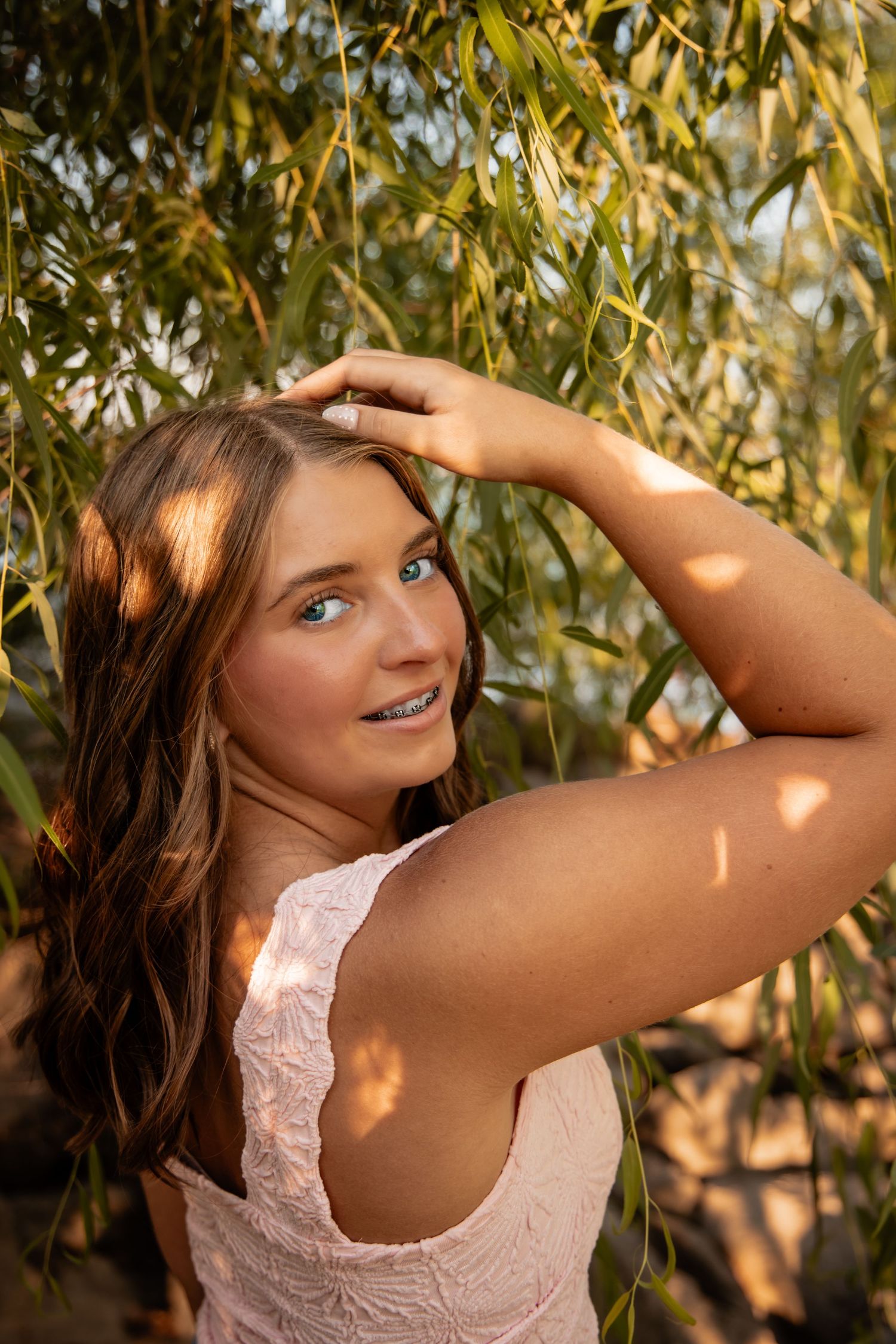 Young woman with blue eyes and braces smiles among willow branches in golden sunlight, wearing a white top.