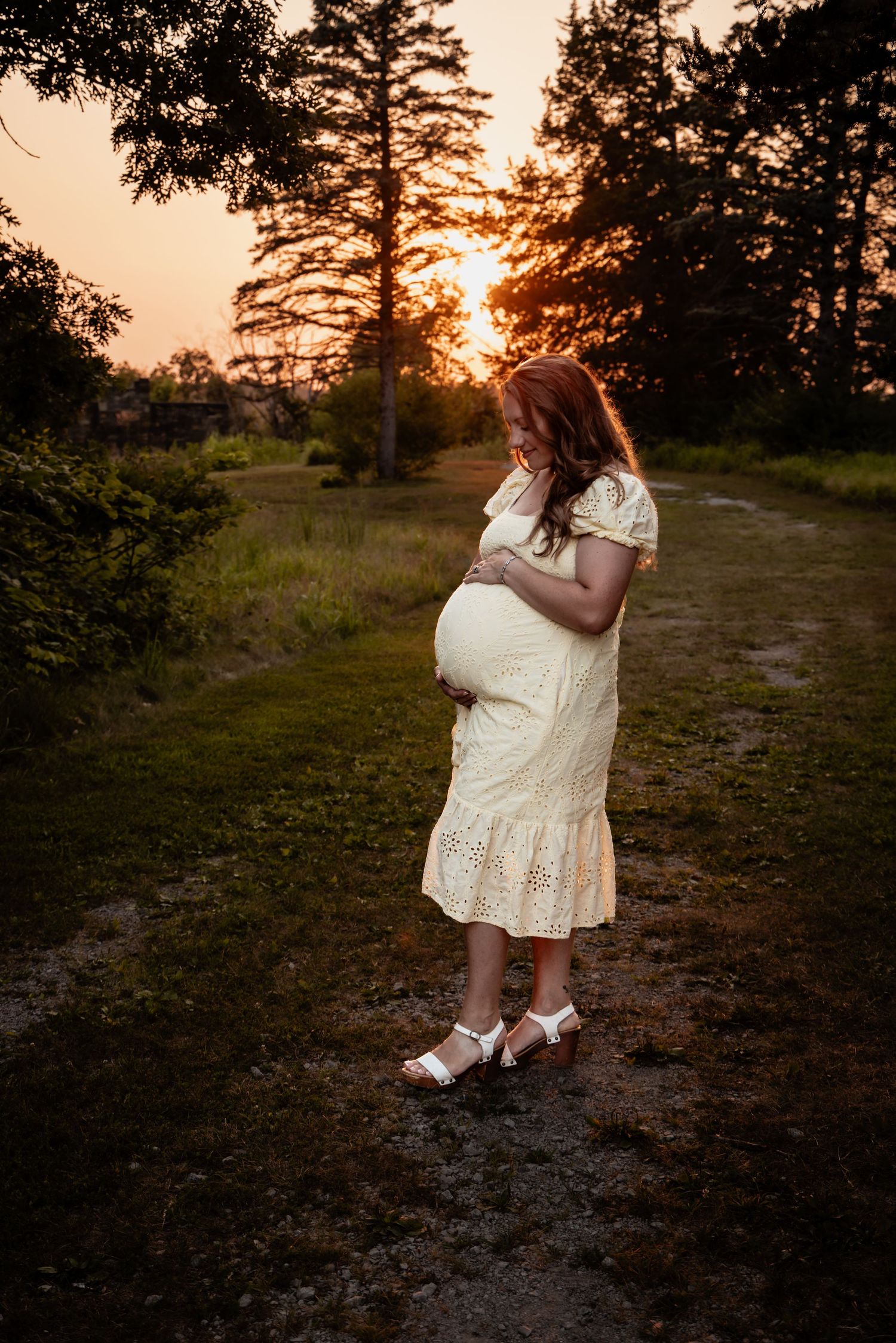 Pregnant woman in white lace dress cradling baby bump at golden sunset in a lush green forest path.