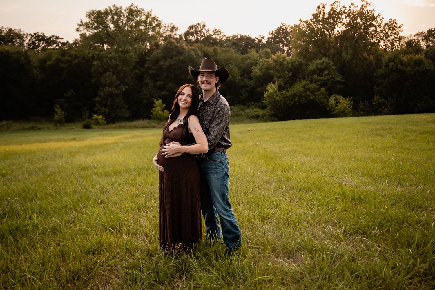 Couple poses in green field at golden hour, pregnant woman in brown gown with man in cowboy hat.