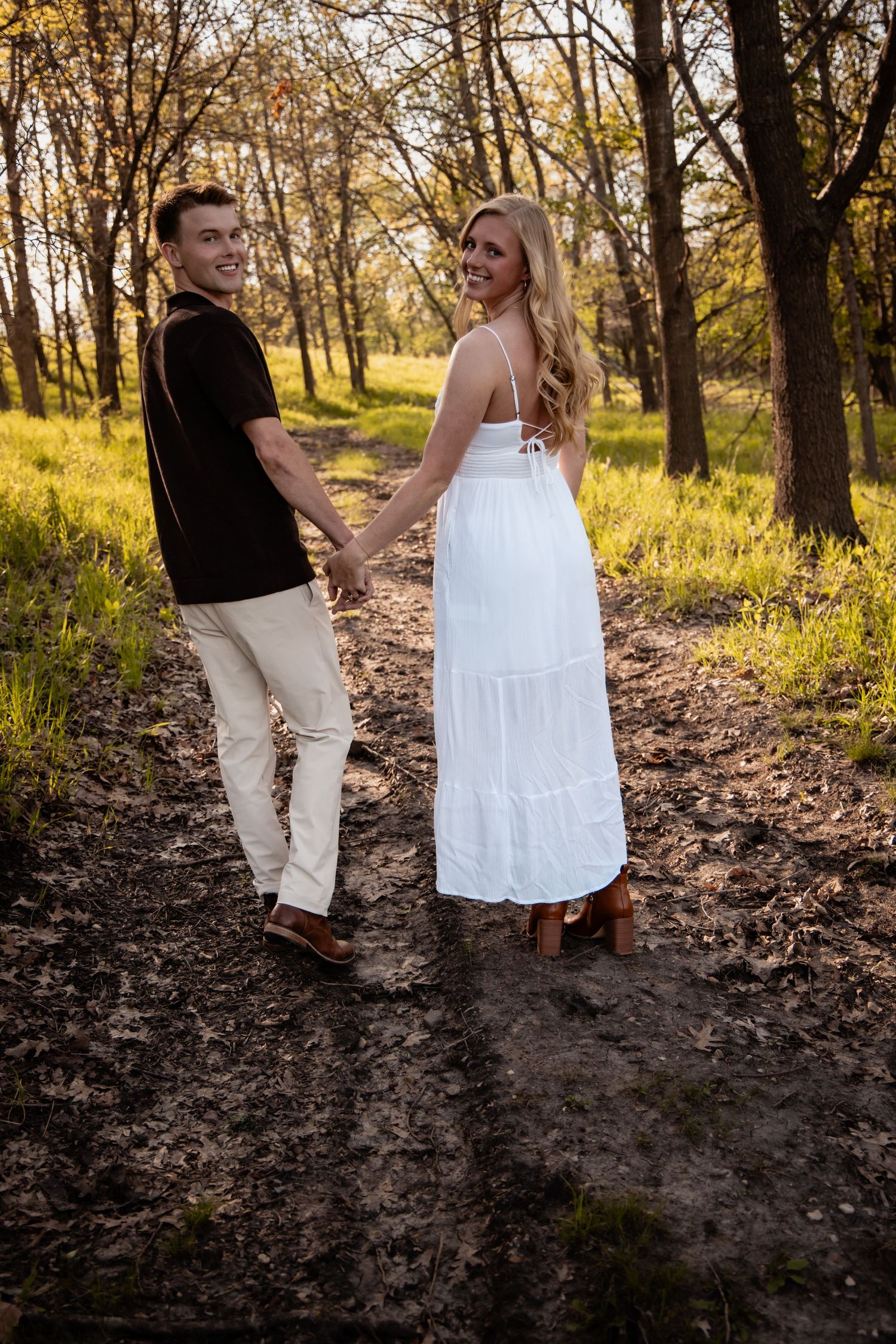 A couple holds hands and smiles while walking along a sunlit forest path, woman in a flowing white dress.