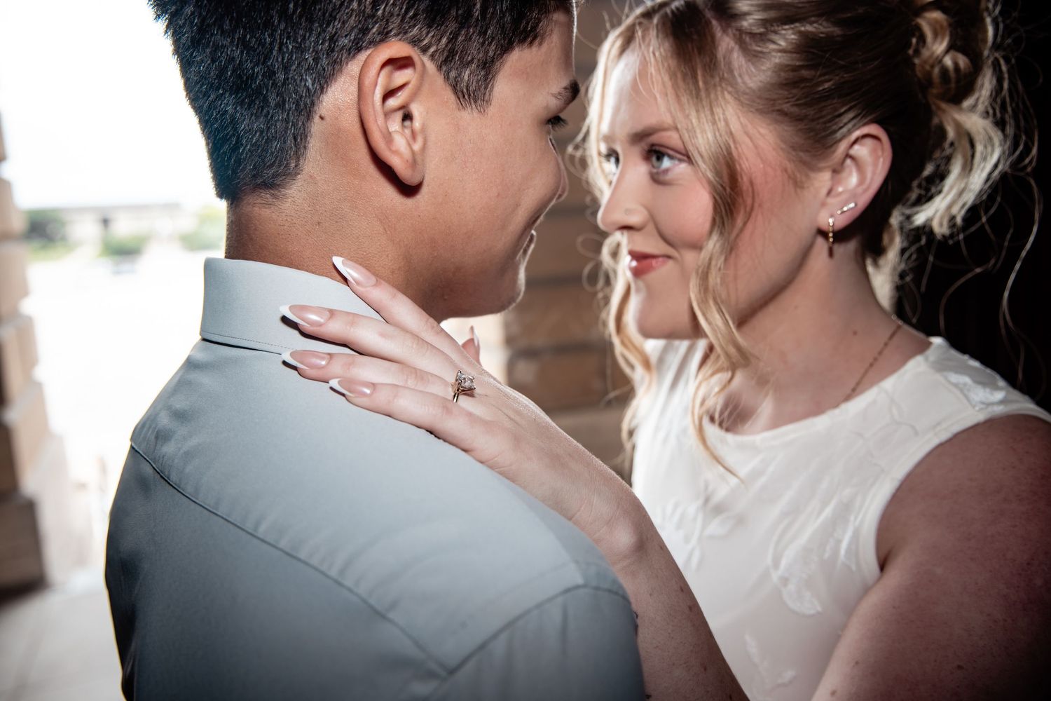 Couple sharing a romantic moment, woman wearing engagement ring rests hands on mans shoulders gazing into his eyes.