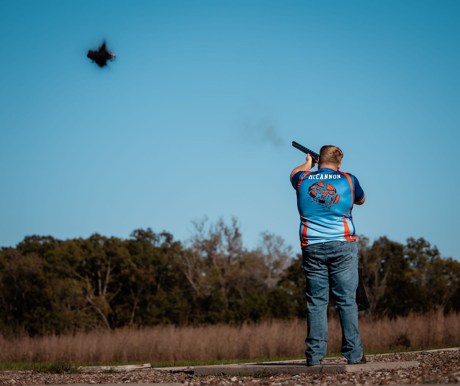 Man in colorful jersey shooting skeet with shotgun outdoors under clear blue sky with trees in background.