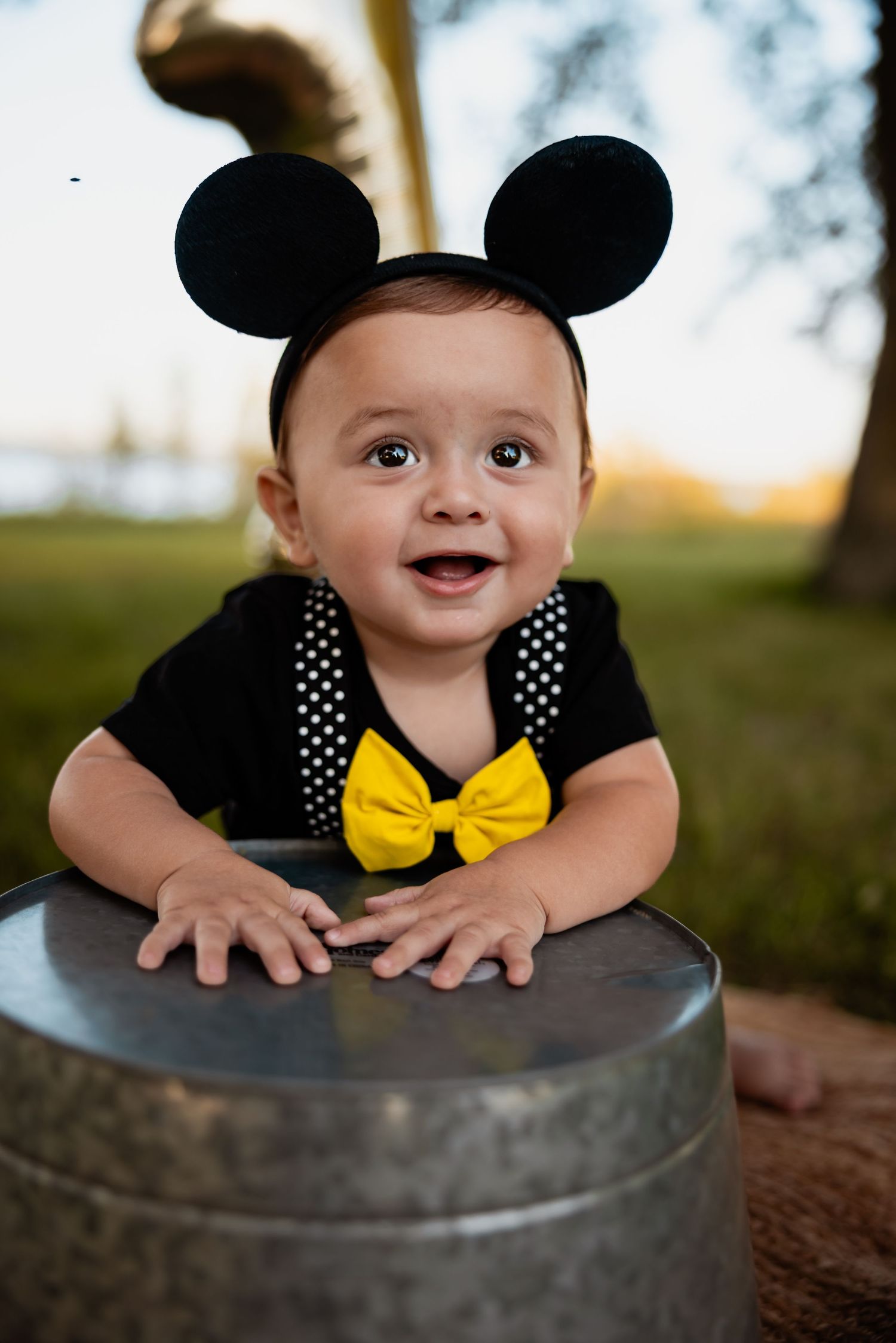 Baby dressed as Mickey Mouse with ears and yellow bow tie, smiling outdoors near a metal bucket.