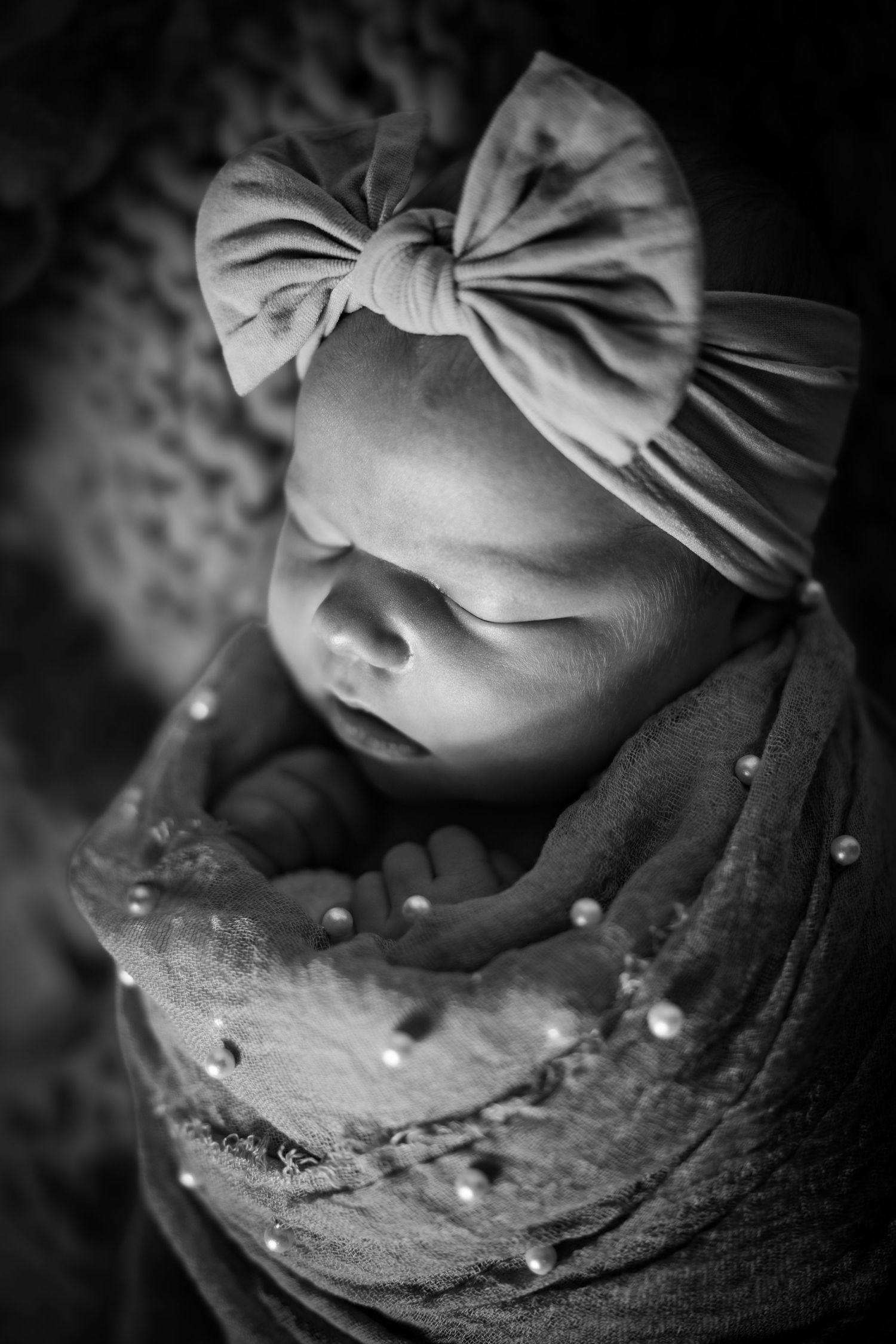 Black and white photo of a sleeping newborn baby wrapped in a pearl-adorned swaddle, wearing a large bow headband.