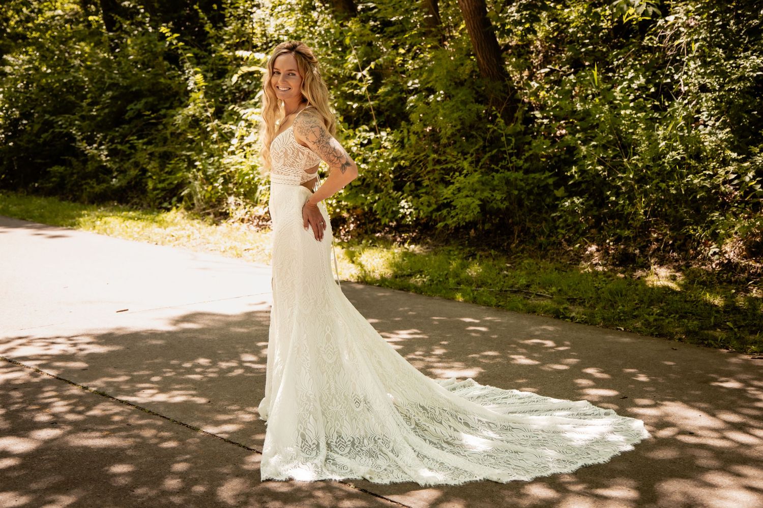 Bride in elegant lace wedding gown with train standing on sunlit path surrounded by lush green trees.