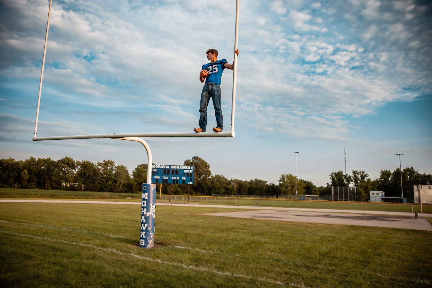 Young football player in blue jersey standing atop a goal post on a green field under a cloudy sky.