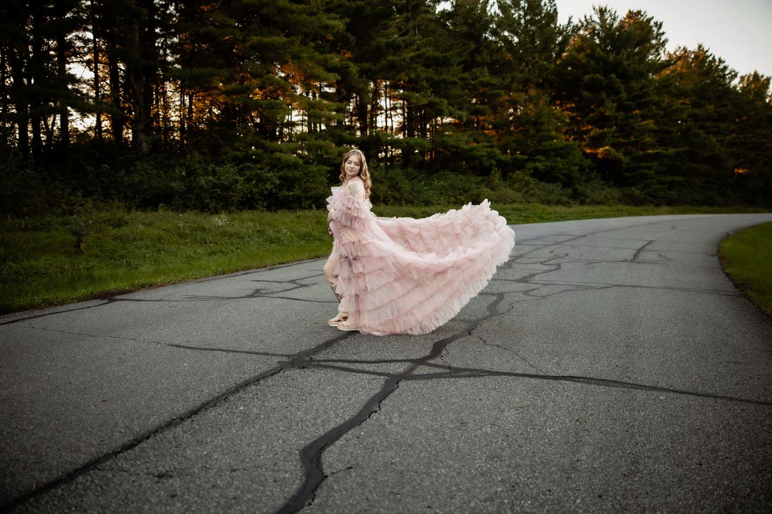 A girl twirls in a flowing pink floral gown on a paved road surrounded by lush green trees at dusk.