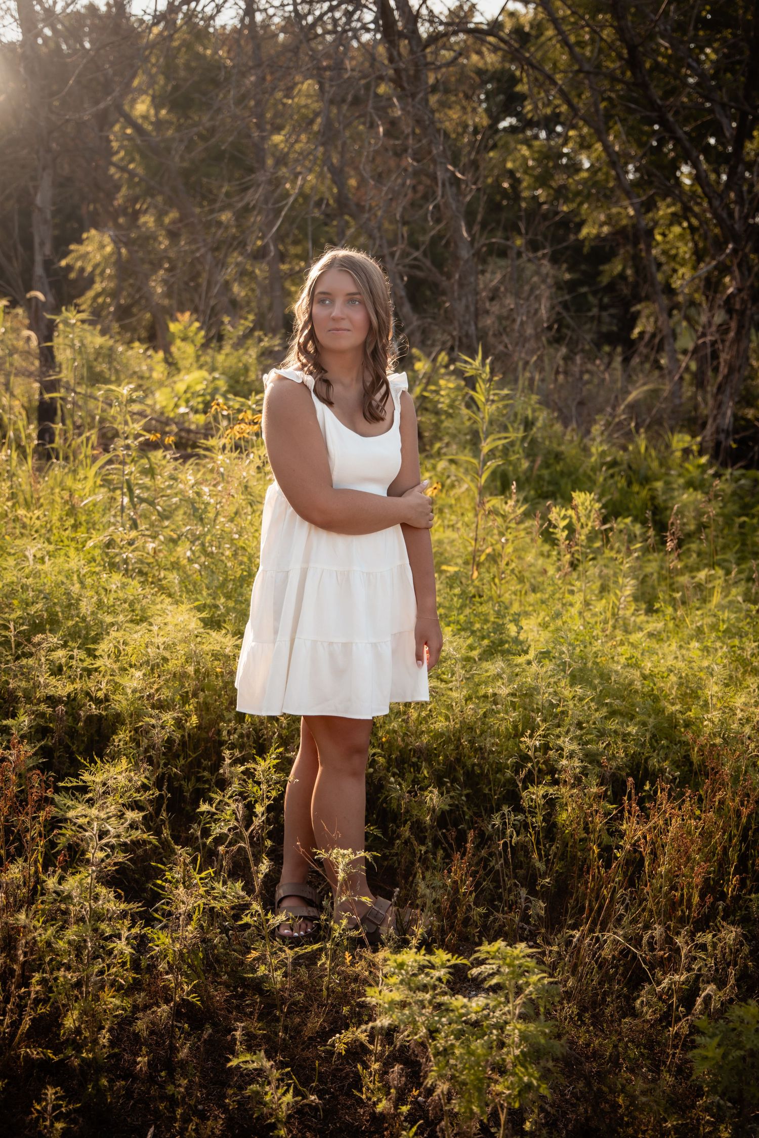 Young woman in white dress standing in sunlit field with lush green foliage and trees in background.