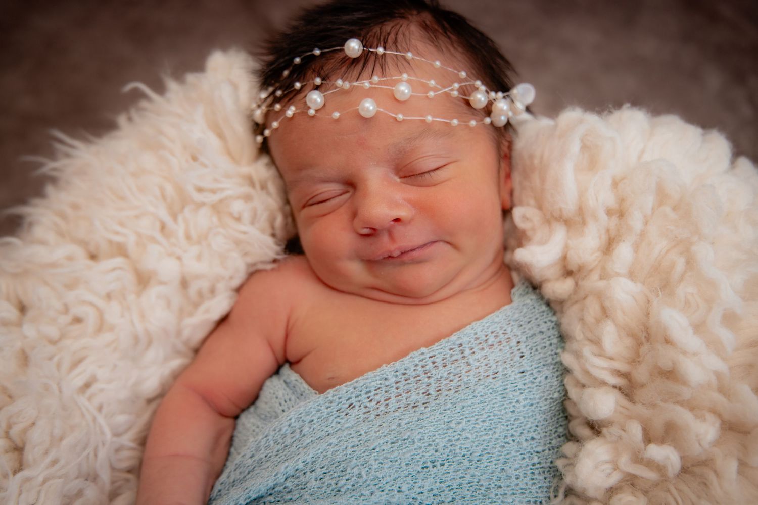Smiling newborn baby wrapped in blue knit blanket wearing pearl headband, nestled in fluffy white fur, professional newborn photo.