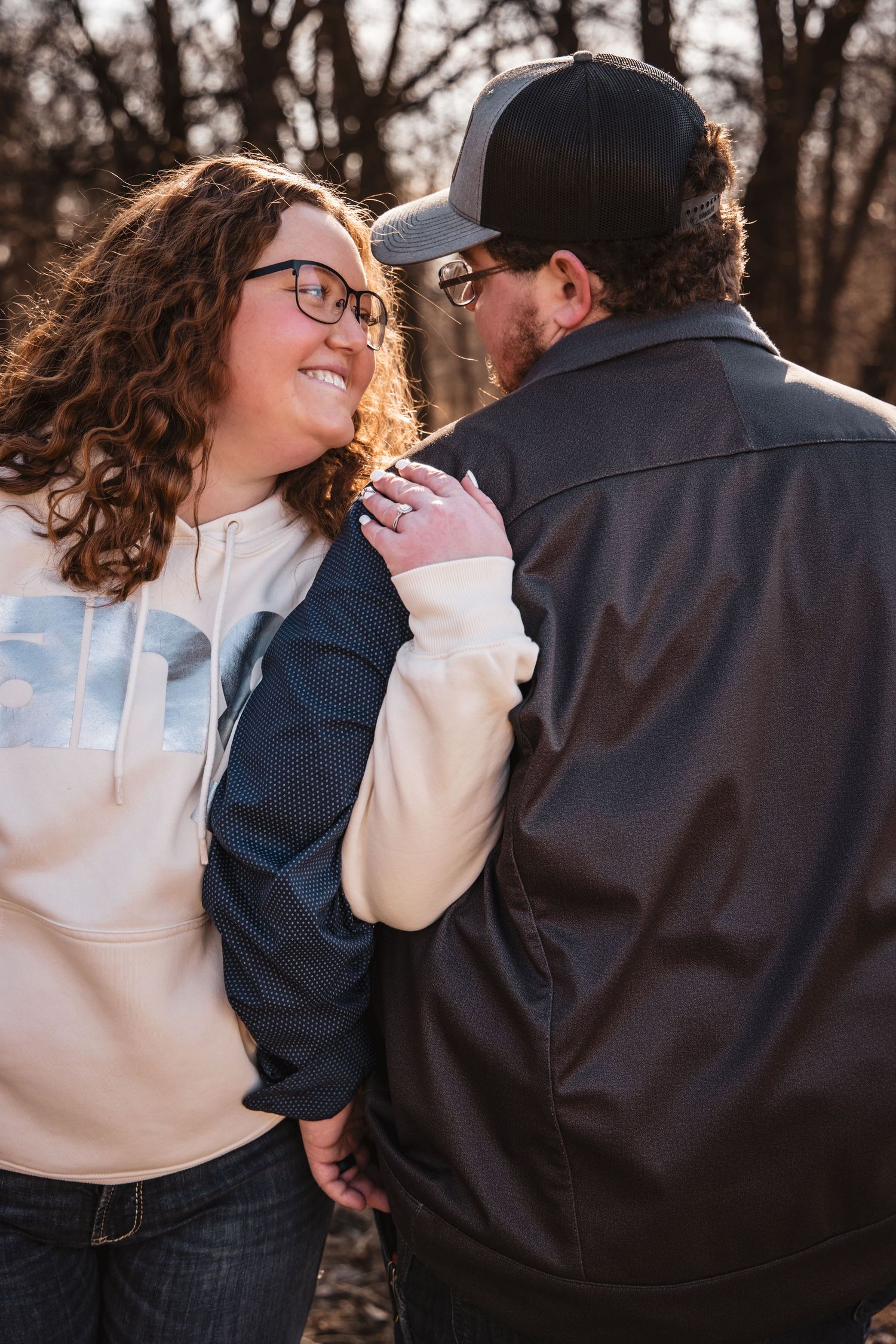 Woman with curly red hair smiles at her partner while holding hands in a sunlit wooded setting.