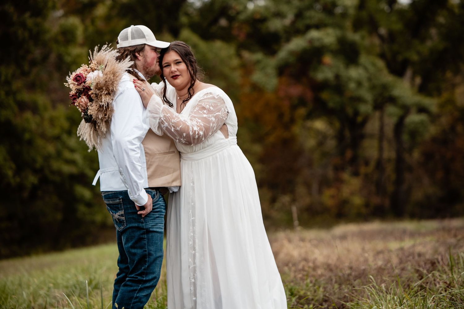 Bride in white lace gown embracing groom in jeans and cap in a scenic autumn field.