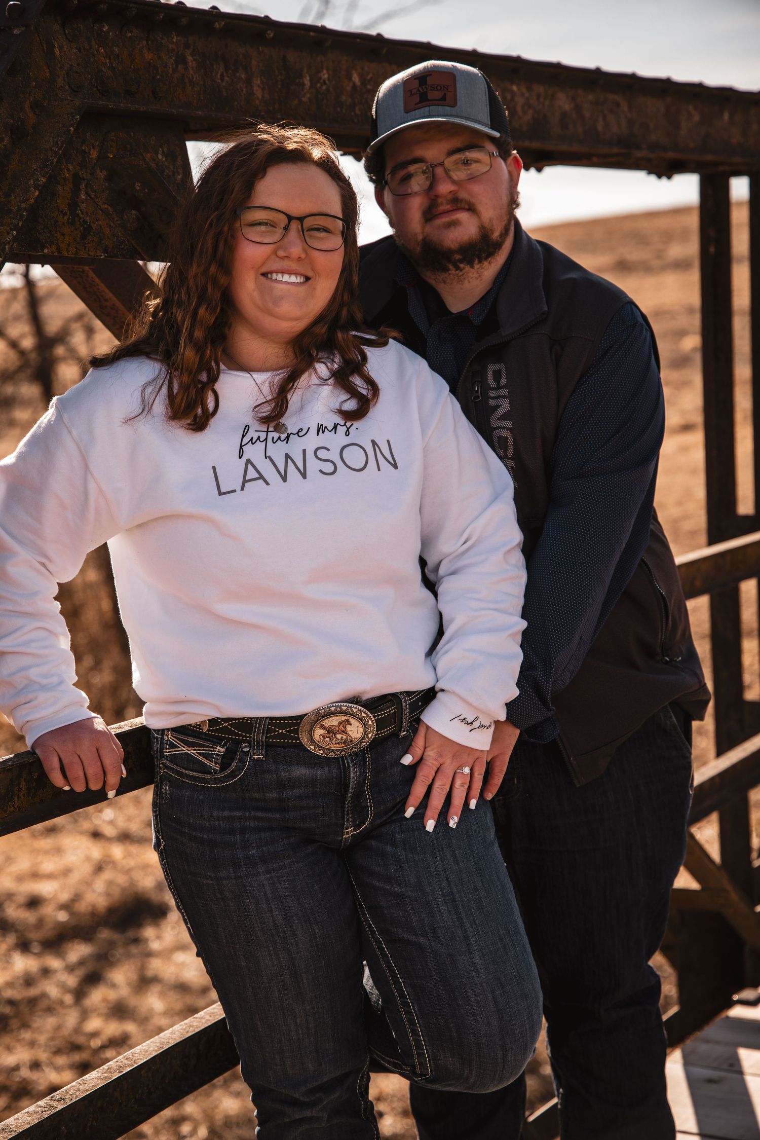 Smiling couple poses on a rustic bridge, woman wearing a Future Mrs. Lawson white sweatshirt.