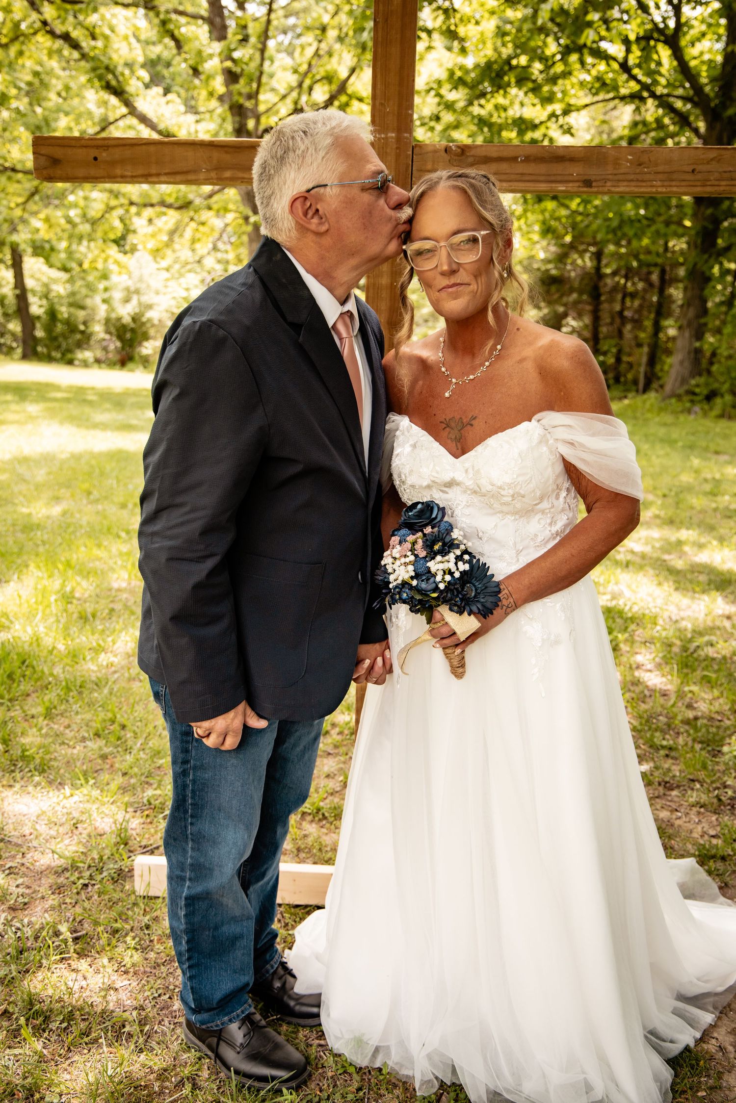 Groom kisses bride on the cheek as she smiles, standing before a rustic wooden cross outdoors.