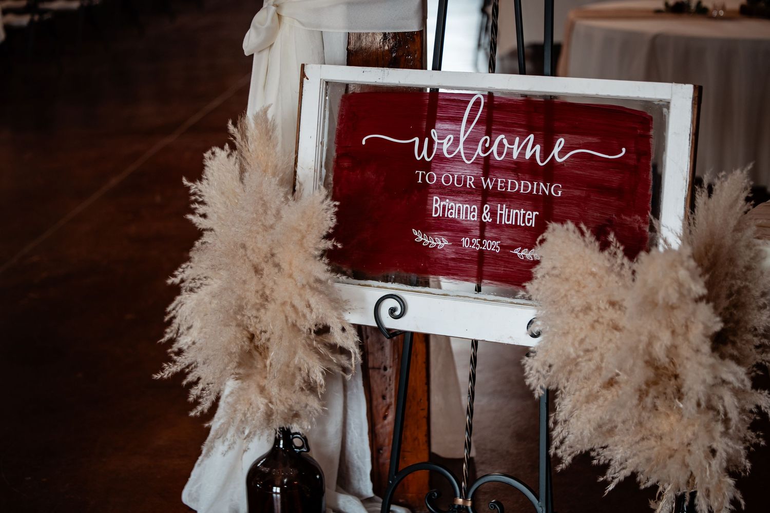 Rustic red welcome wedding sign with white script lettering flanked by pampas grass decor.
