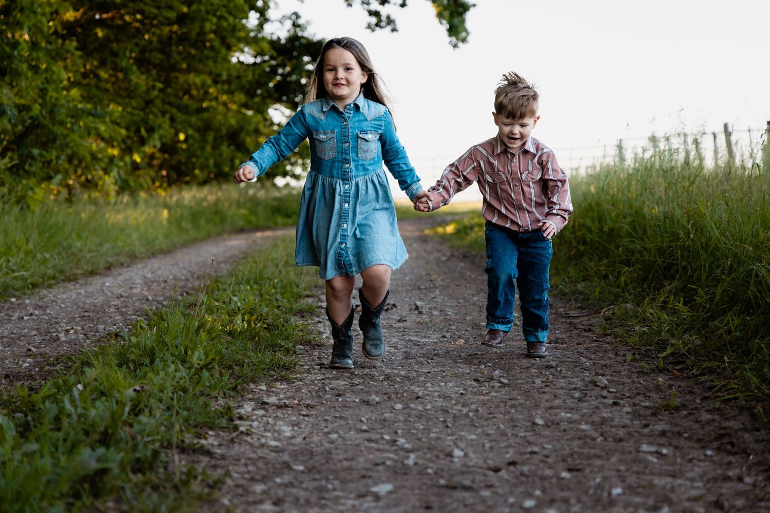 A young girl in a denim dress and boy in a plaid shirt hold hands walking down a dirt path.