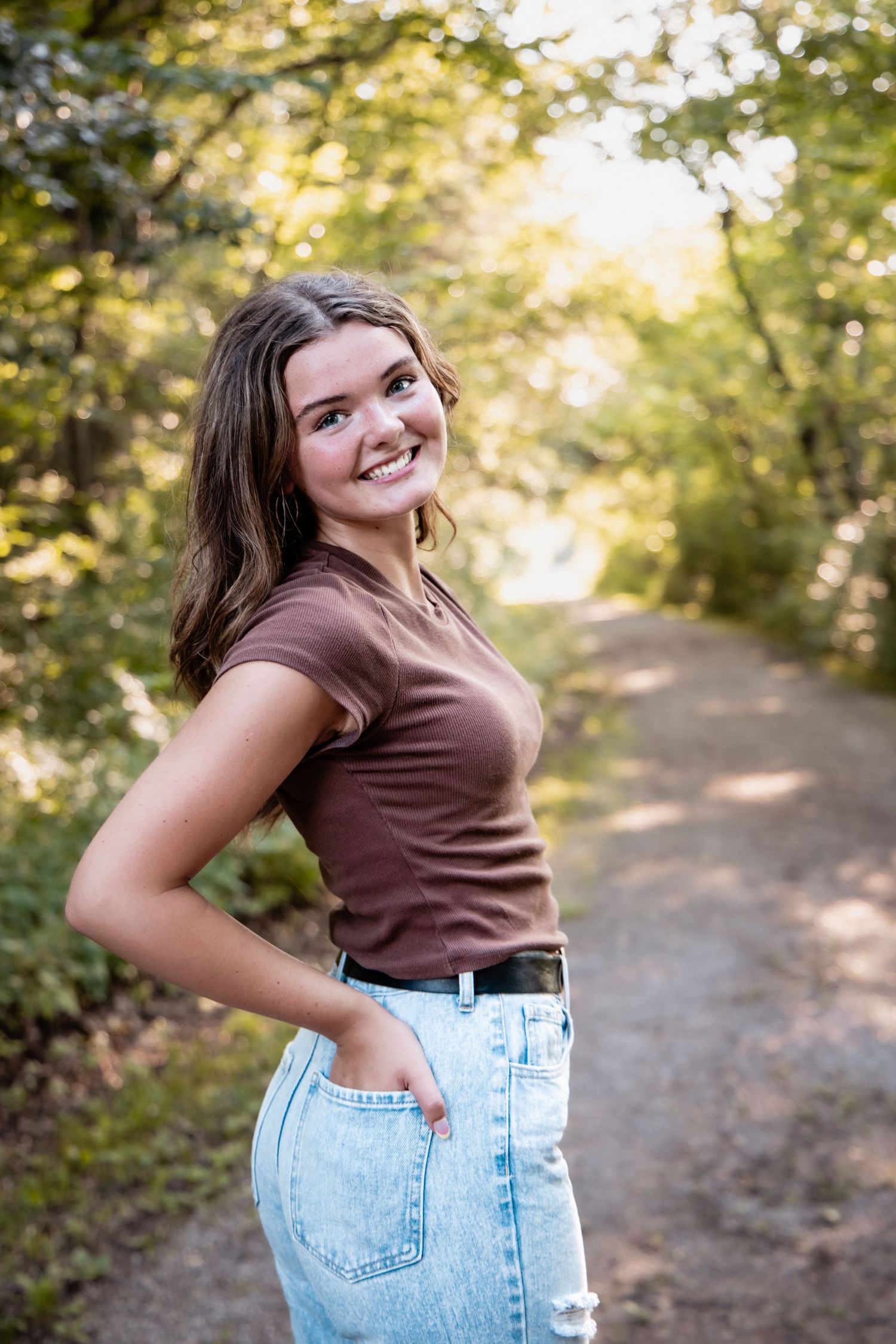 Smiling teenage girl in brown top and jeans poses on a tree-lined dirt path in golden sunlight.