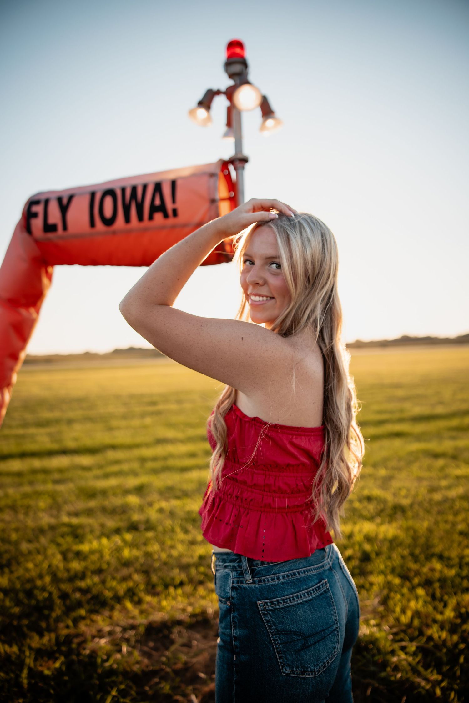 Blonde woman in red top and jeans poses in front of a Fly Iowa sign on a sunny green field.