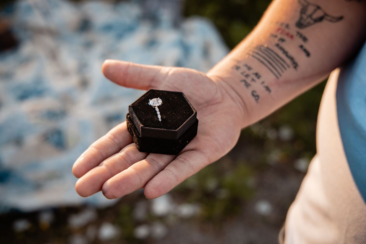 Tattooed hand holding a black octagonal ring box with a diamond engagement ring inside, outdoors.