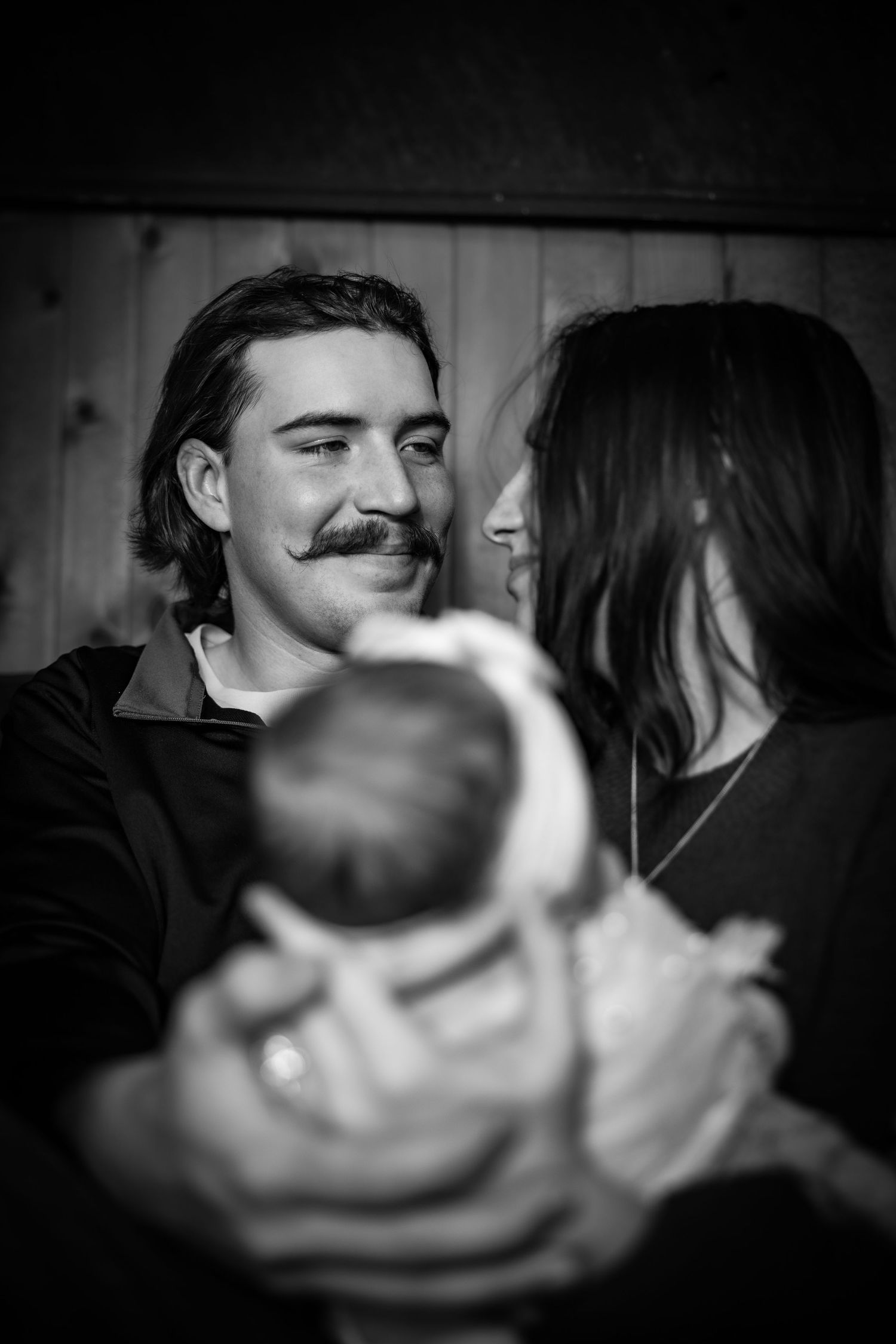 Black and white photo of a smiling man with mustache and woman gazing at each other while holding a newborn baby.