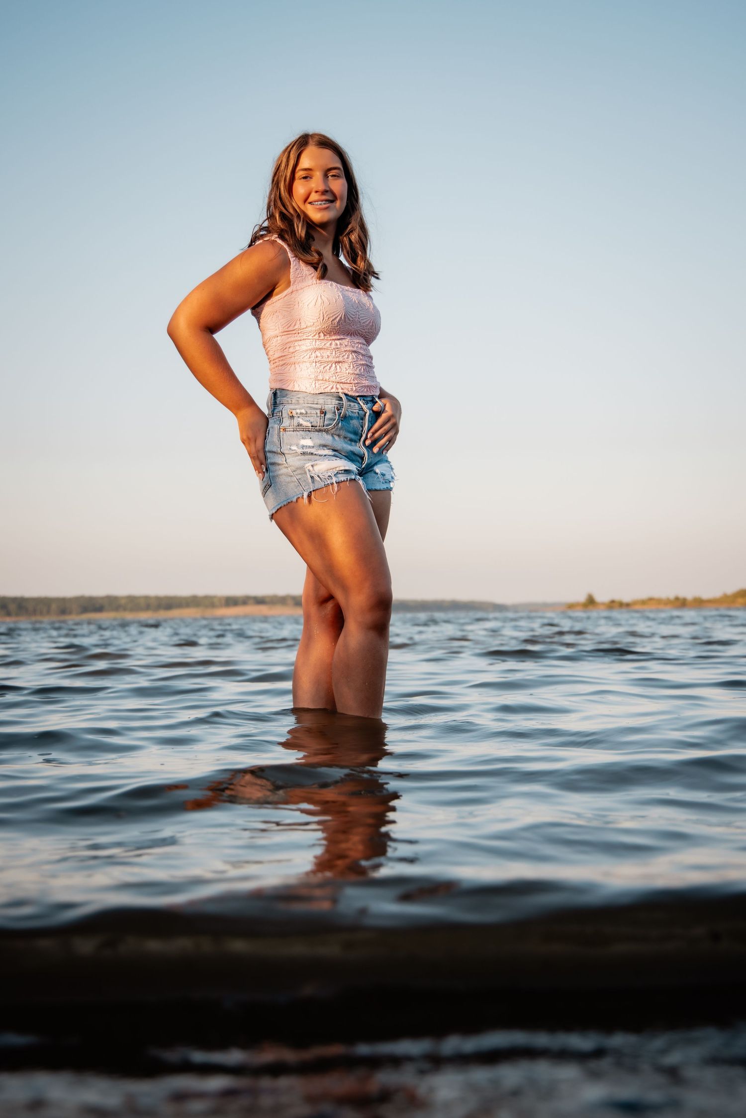 Young woman in white tank top and denim shorts standing in shallow lake water at sunset.