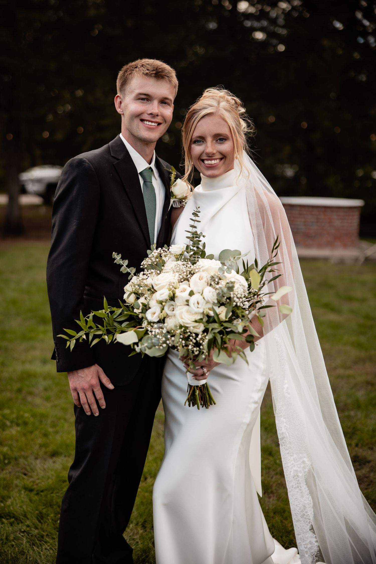 Smiling bride and groom pose outdoors, bride holding white floral bouquet with greenery.