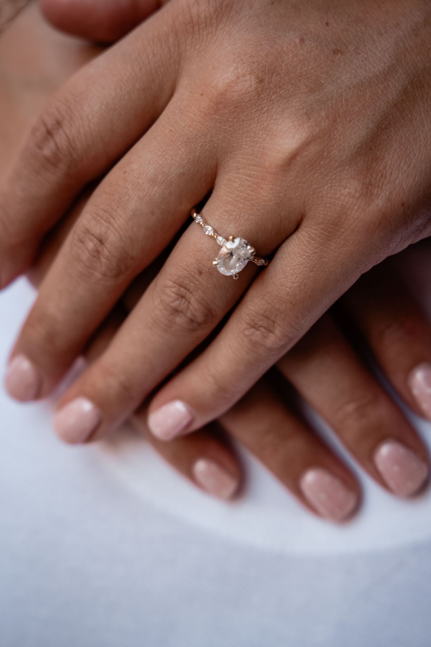 Close-up of a delicate diamond solitaire engagement ring on a woman's finger with nude pink nails.