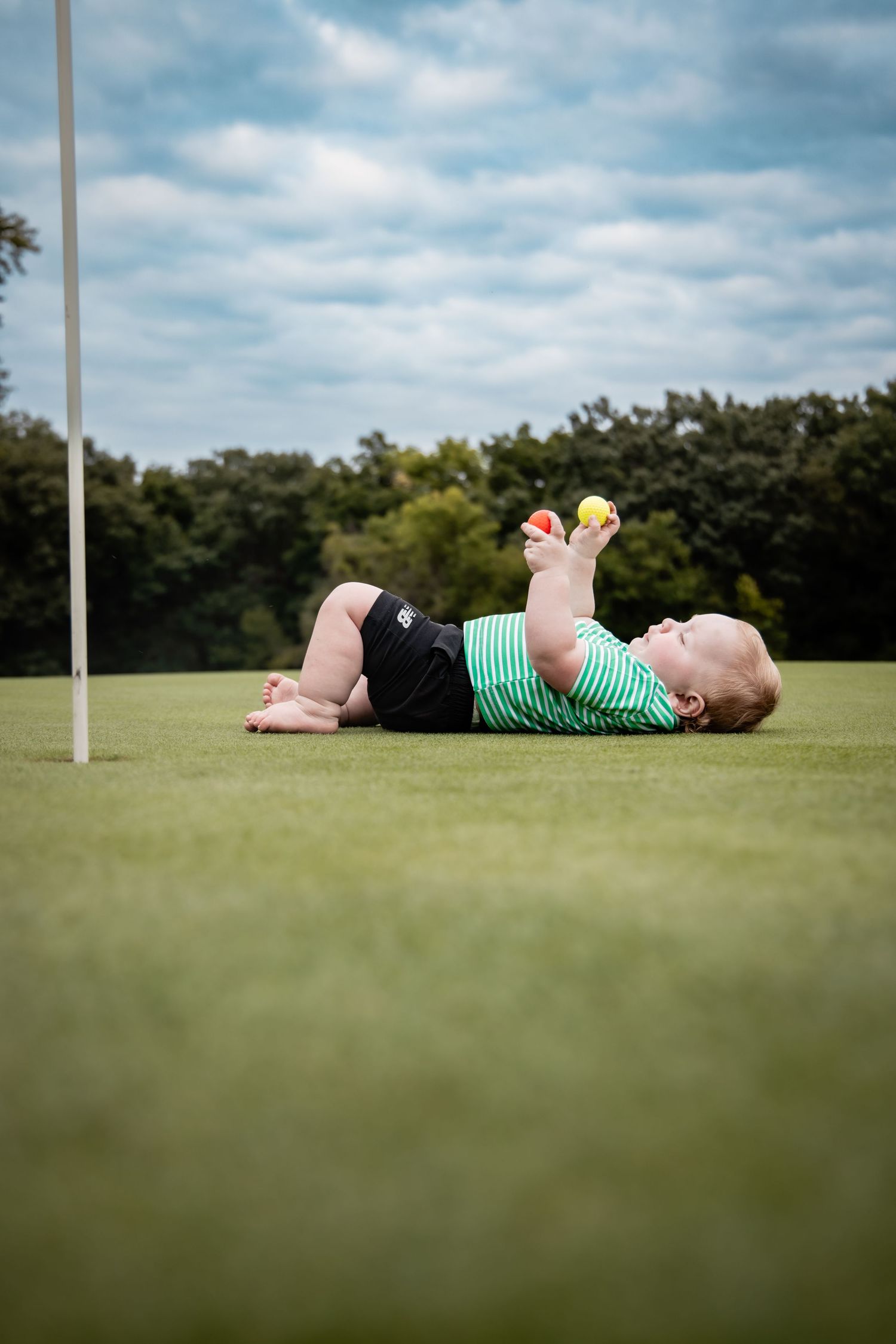 Baby lying on green golf course grass, holding small colorful balls up in the air, wearing striped onesie.