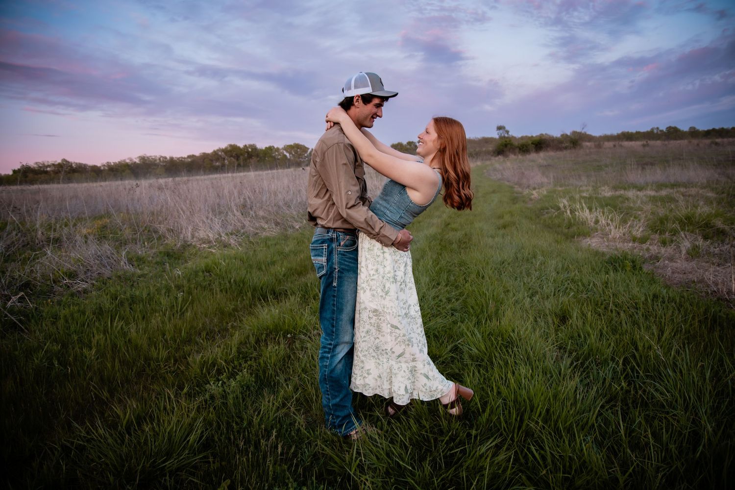 Couple embracing romantically in a green field under a dramatic purple sunset sky.